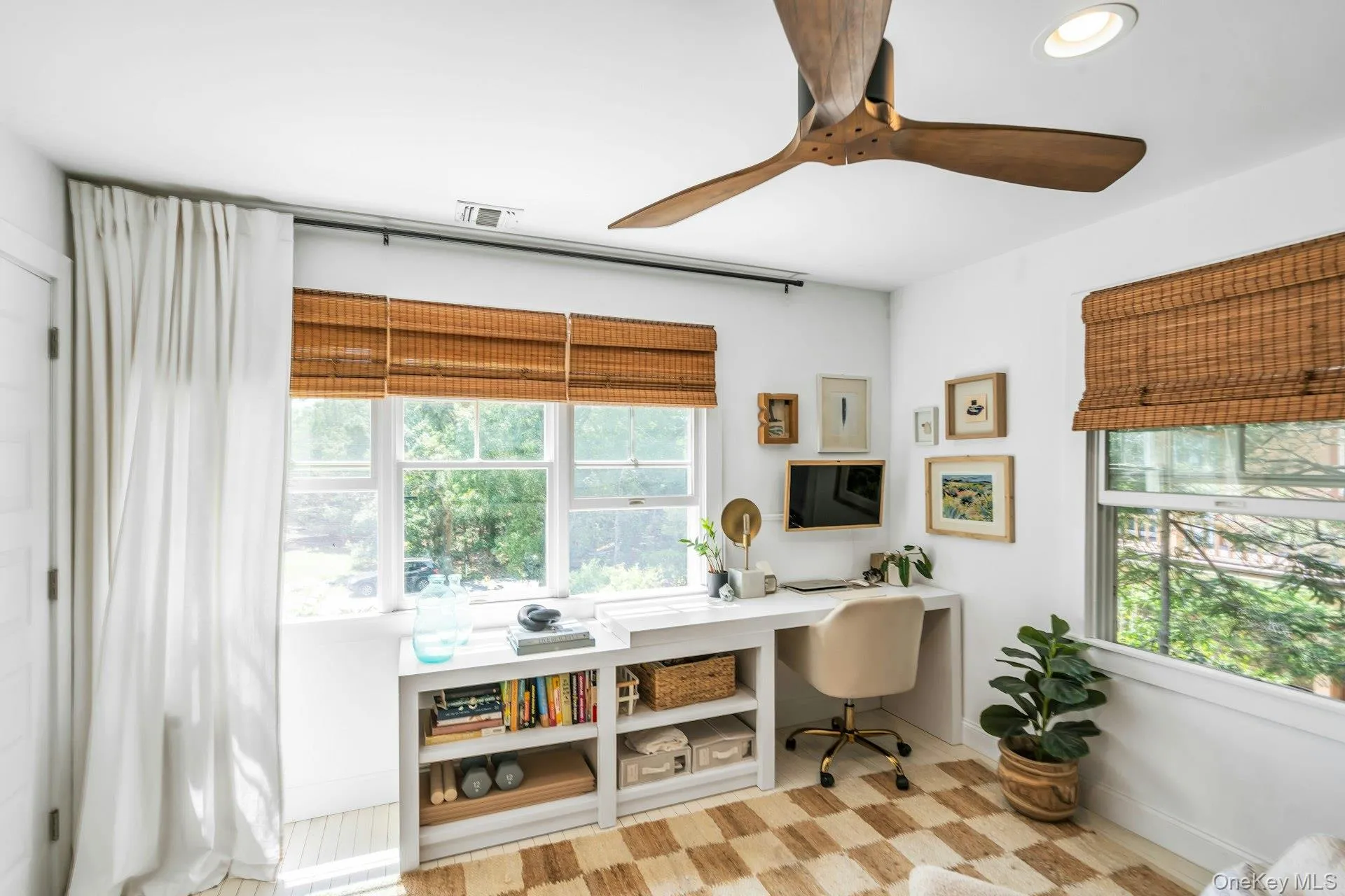 Bright room featuring multiple windows with natural woven shades, a built-in desk with integrated shelving, and a ceiling fan with wood-finish blades Bright room featuring multiple windows with natural woven shades, a built-in desk with integrated shelving, and a ceiling fan with wood-finish blades