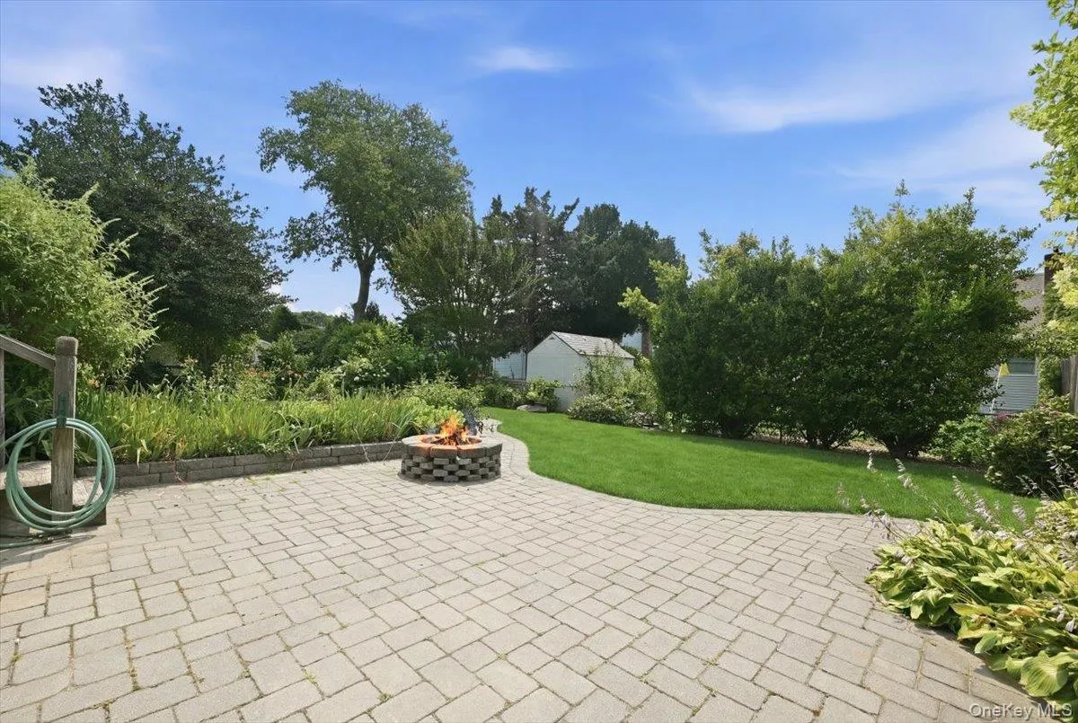 Spacious backyard featuring a paver patio with a fire pit, manicured lawn, lush landscaping, and mature trees Spacious backyard featuring a paver patio with a fire pit, manicured lawn, lush landscaping, and mature trees