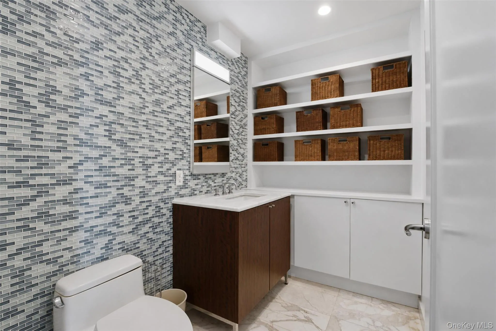 Powder room featuring a full wall of mosaic tile, a wood-finish vanity with a white countertop, and extensive built-in shelving Powder room featuring a full wall of mosaic tile, a wood-finish vanity with a white countertop, and extensive built-in shelving