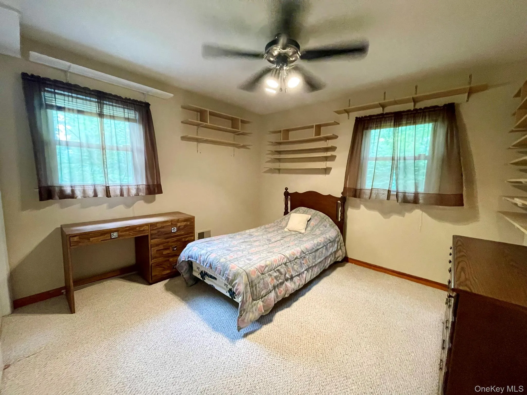 Carpeted room featuring a ceiling fan with integrated lighting, two windows with brown sheer curtains, and multiple wall-mounted shelving units Carpeted room featuring a ceiling fan with integrated lighting, two windows with brown sheer curtains, and multiple wall-mounted shelving units