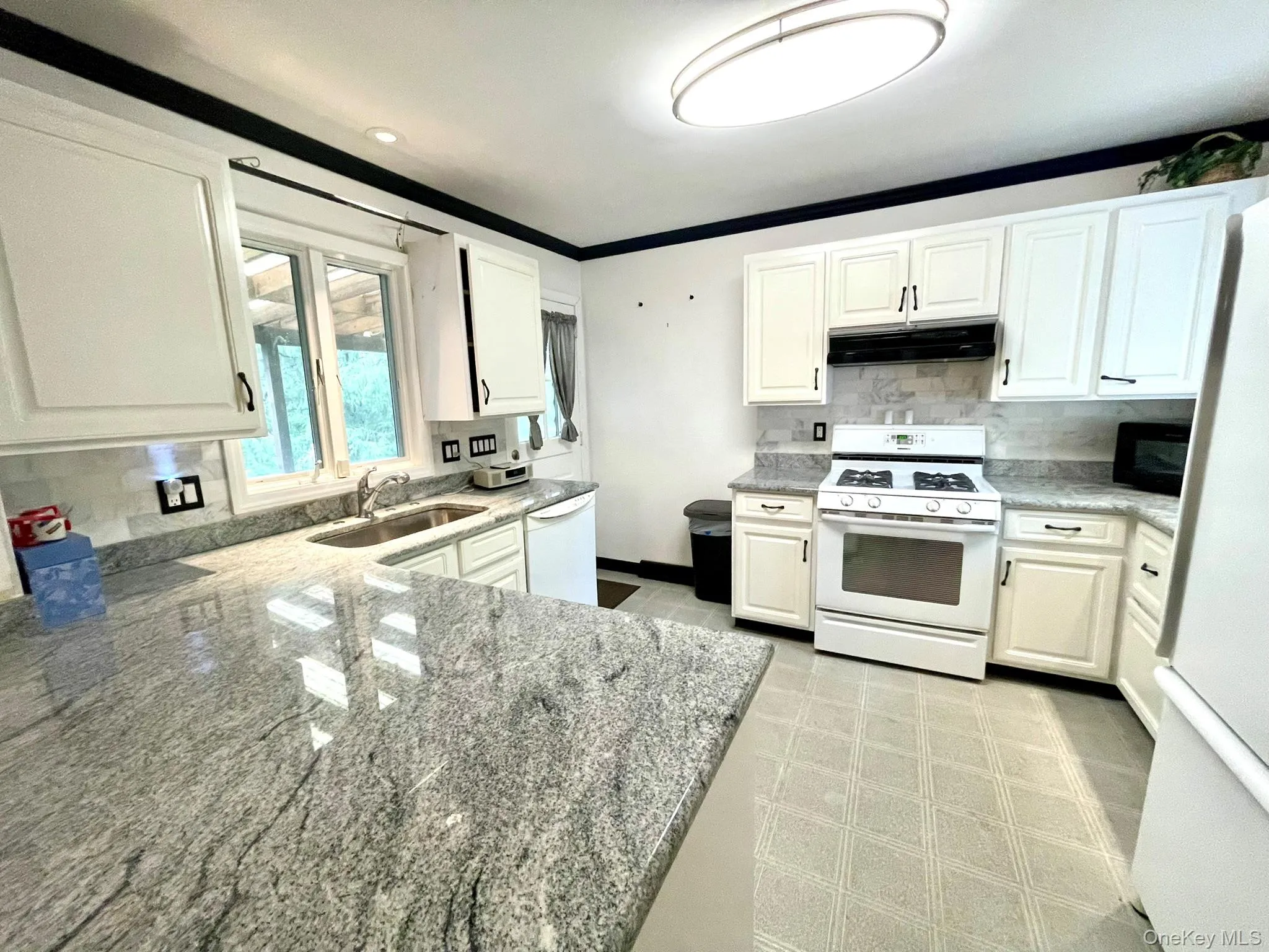 Kitchen featuring light-toned countertops, white cabinetry with dark hardware, a white range, a stainless steel sink, and a tiled backsplash Kitchen featuring light-toned countertops, white cabinetry with dark hardware, a white range, a stainless steel sink, and a tiled backsplash