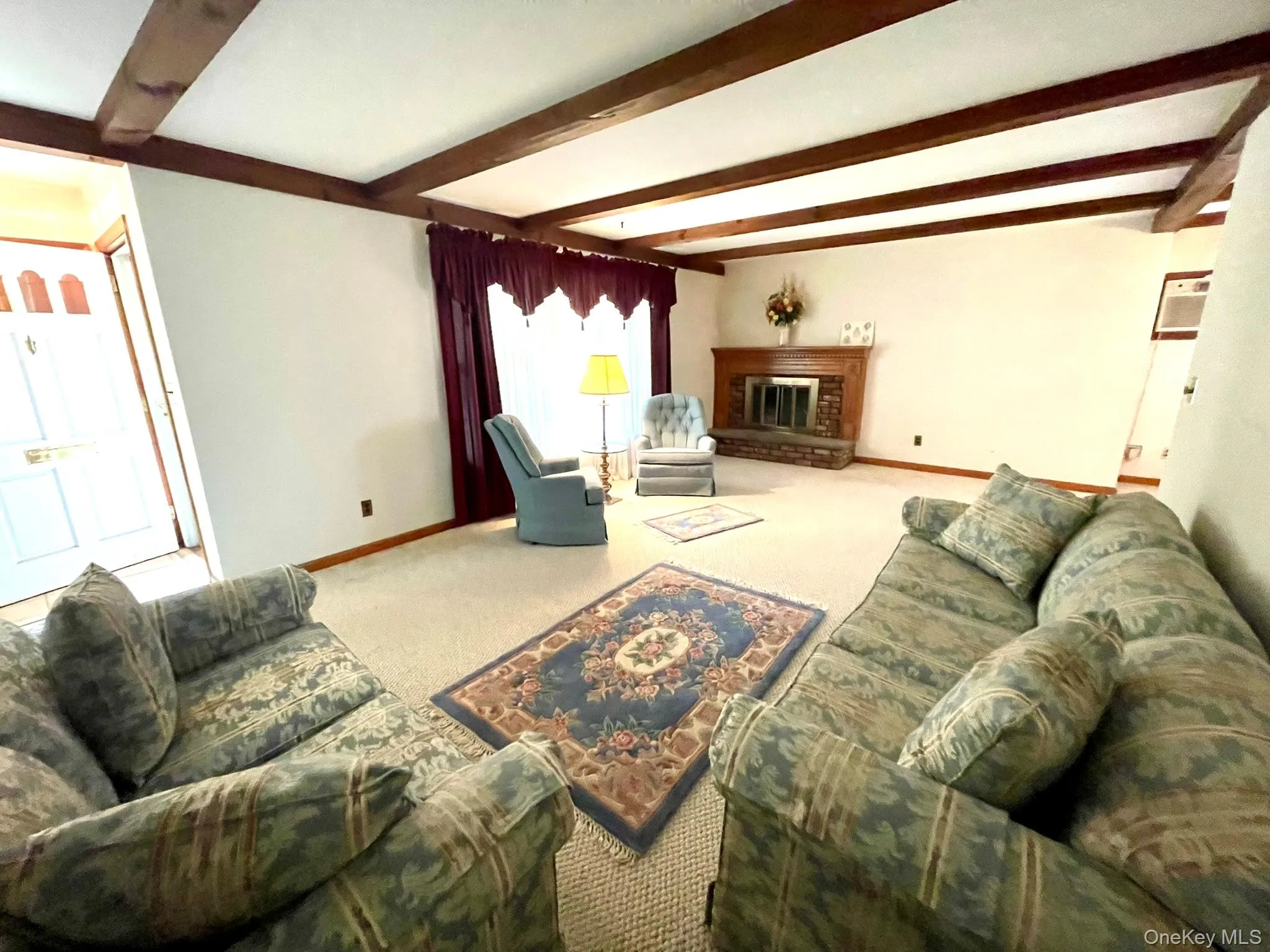 Living area featuring exposed wood beams, a prominent fireplace with a wood mantle and stone hearth, and light-colored carpeting Living area featuring exposed wood beams, a prominent fireplace with a wood mantle and stone hearth, and light-colored carpeting