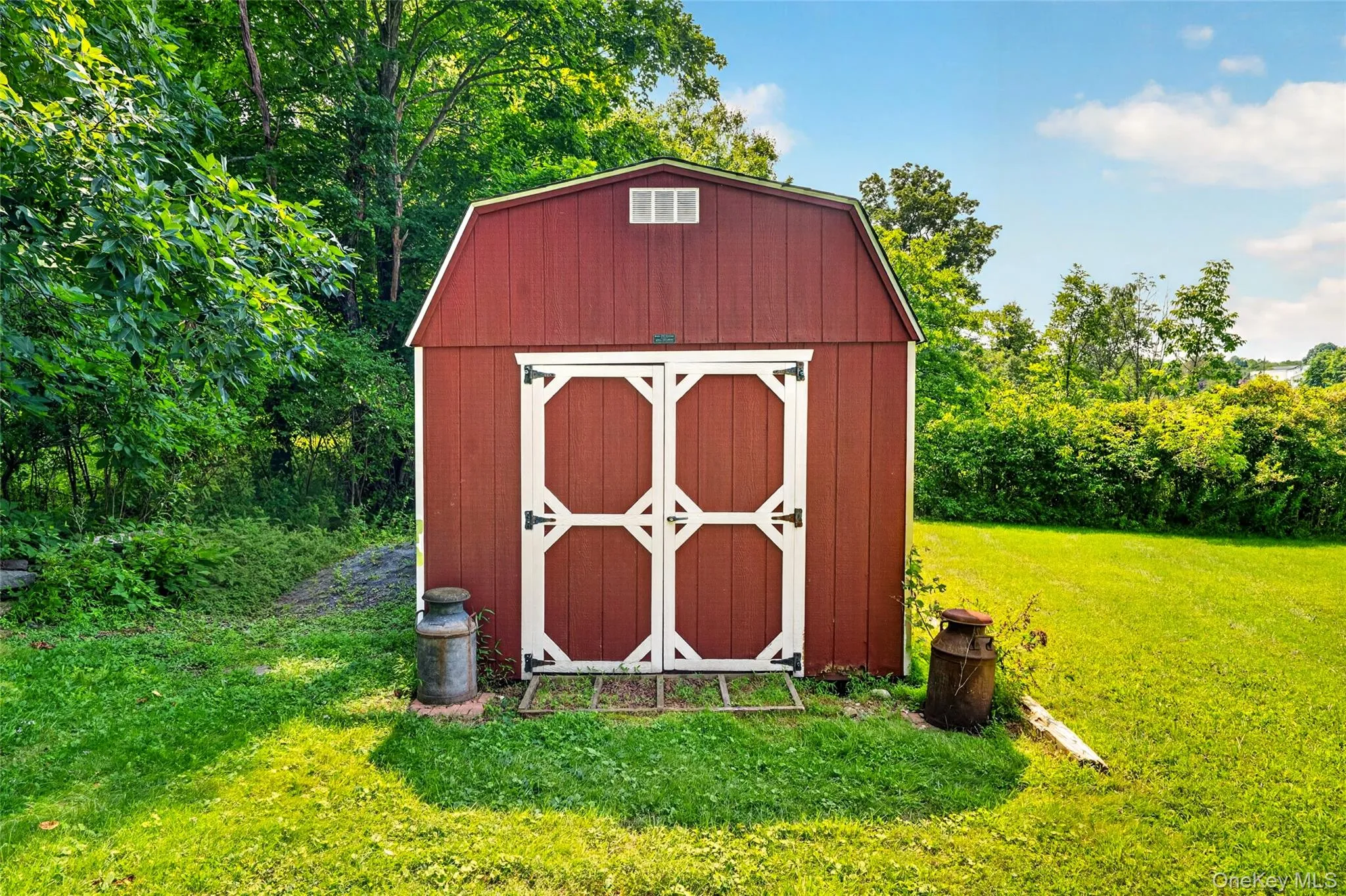 Red gambrel-style shed with white double doors, set on a green lawn with a wooded backdrop Red gambrel-style shed with white double doors, set on a green lawn with a wooded backdrop