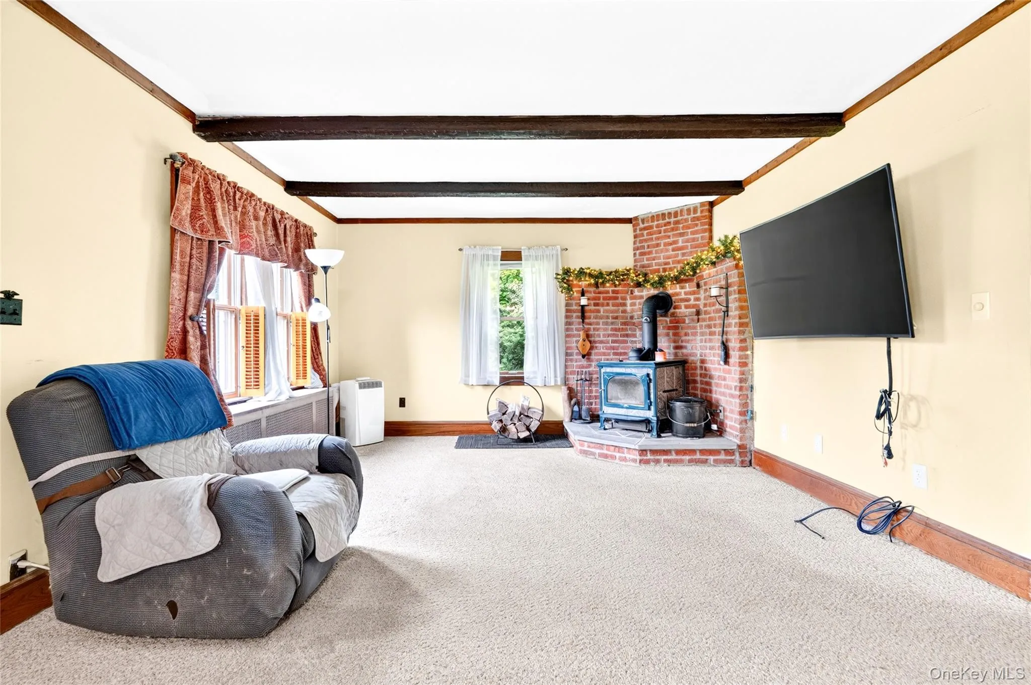 Exposed ceiling beams, brick corner fireplace, wood-burning stove, wall-mounted television, and light-colored carpeting Exposed ceiling beams, brick corner fireplace, wood-burning stove, wall-mounted television, and light-colored carpeting