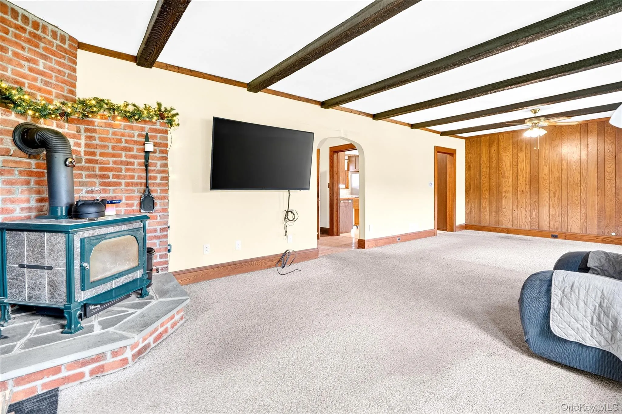 Exposed wood beam ceiling, brick fireplace with stone hearth, wood-burning stove, wood paneling, and ceiling fan Exposed wood beam ceiling, brick fireplace with stone hearth, wood-burning stove, wood paneling, and ceiling fan