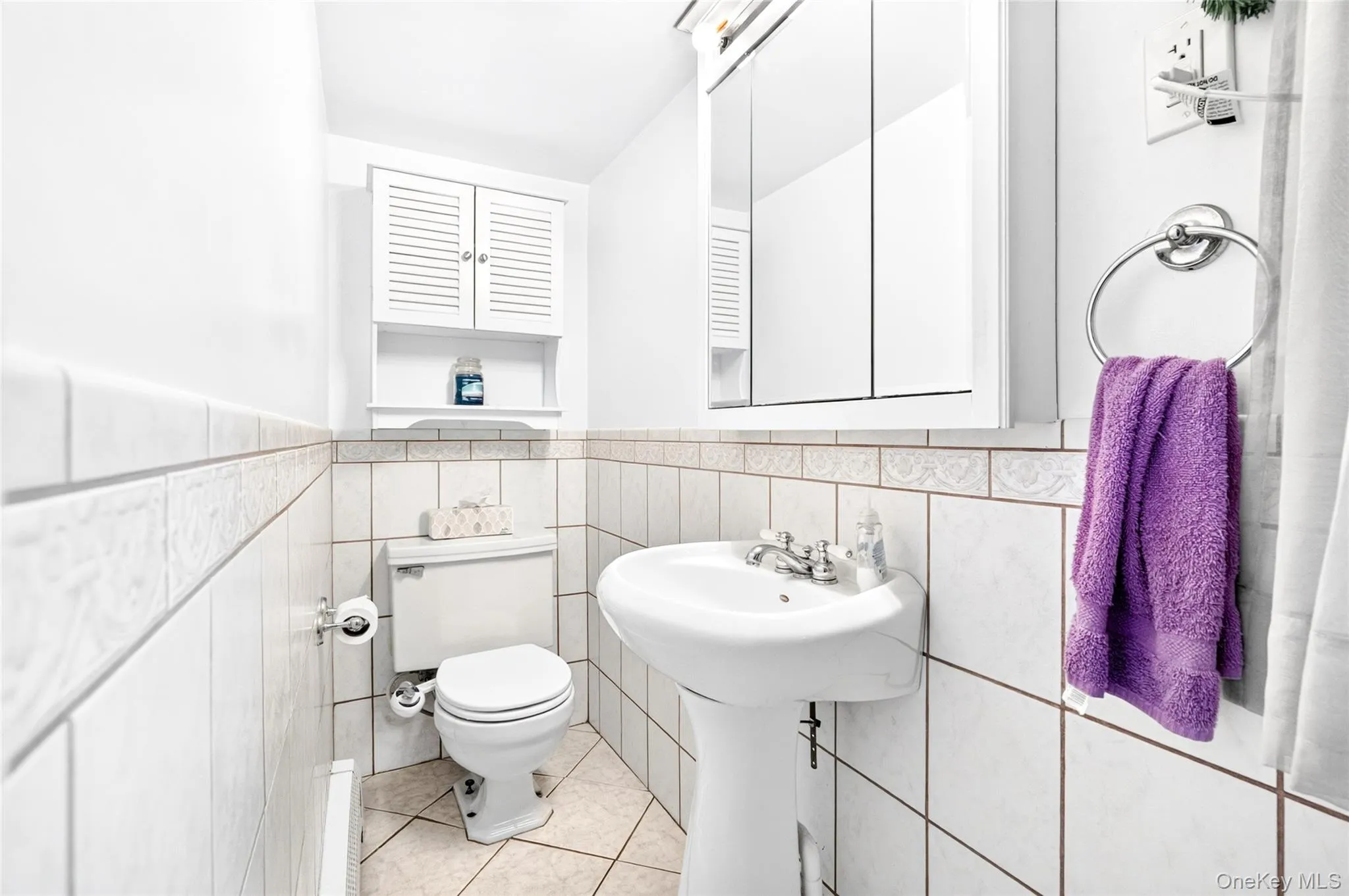 White tiled walls with decorative trim, white pedestal sink, white toilet, wall-mounted white medicine cabinet, and wall-mounted white louvered cabinet White tiled walls with decorative trim, white pedestal sink, white toilet, wall-mounted white medicine cabinet, and wall-mounted white louvered cabinet