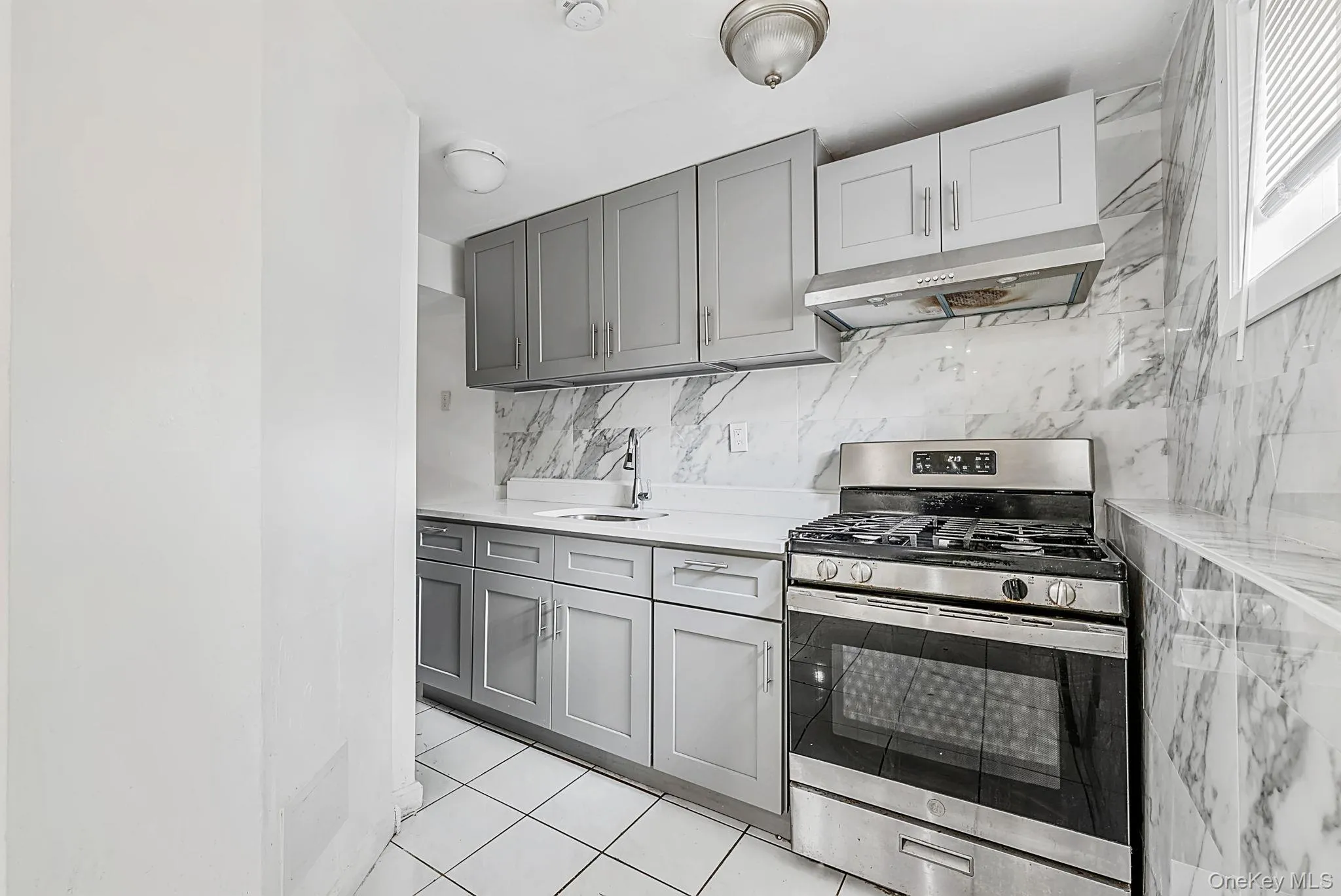 Kitchen featuring grey cabinetry, stainless steel range with overhead ventilation, undermount sink, marble-look backsplash, and white tiled flooring Kitchen featuring grey cabinetry, stainless steel range with overhead ventilation, undermount sink, marble-look backsplash, and white tiled flooring