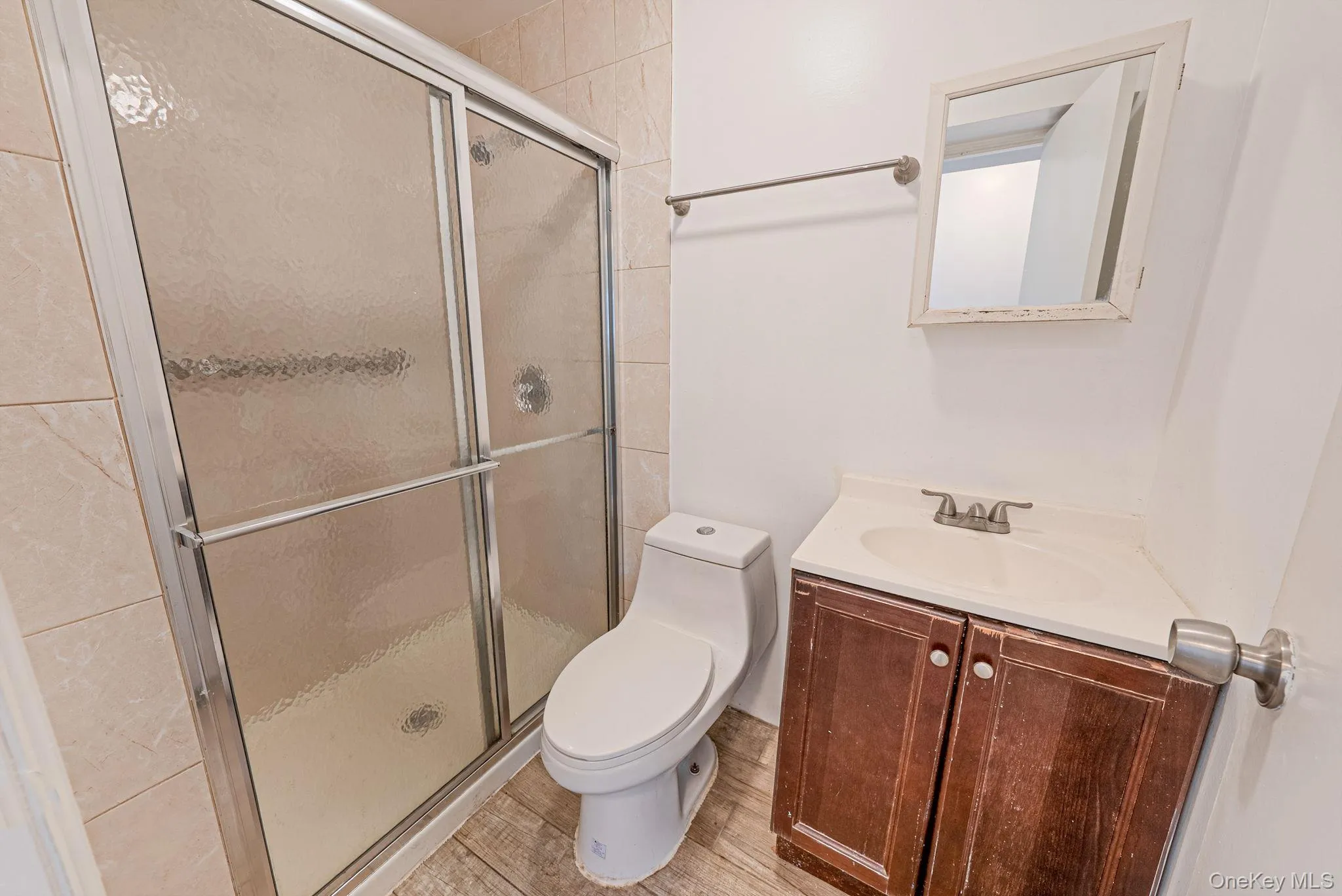 Bathroom featuring a sliding glass shower enclosure, a vanity with a white countertop and dark wood-finish cabinetry, a white toilet, and wood-finish flooring Bathroom featuring a sliding glass shower enclosure, a vanity with a white countertop and dark wood-finish cabinetry, a white toilet, and wood-finish flooring