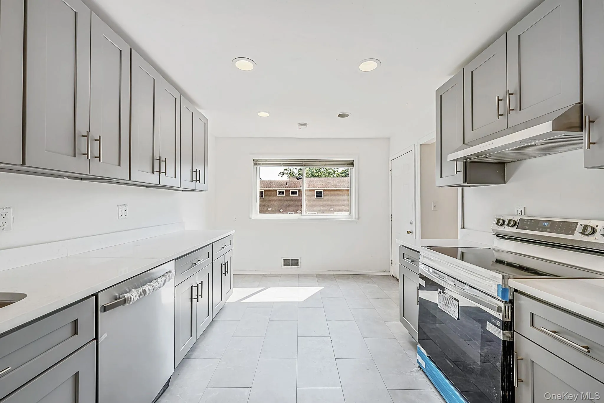Modern kitchen featuring an integrated electric range with an overhead vent hood, stainless steel dishwasher, extensive gray cabinetry with bar pulls, and a window providing natural light Modern kitchen featuring an integrated electric range with an overhead vent hood, stainless steel dishwasher, extensive gray cabinetry with bar pulls, and a window providing natural light