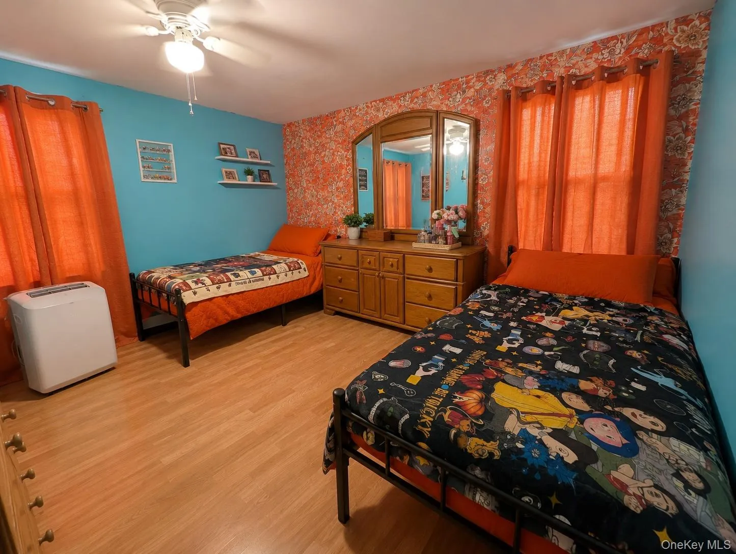 Spacious bedroom featuring wood-finish flooring, a ceiling fan with integrated lighting, and a prominent feature wall with patterned wallpaper Spacious bedroom featuring wood-finish flooring, a ceiling fan with integrated lighting, and a prominent feature wall with patterned wallpaper