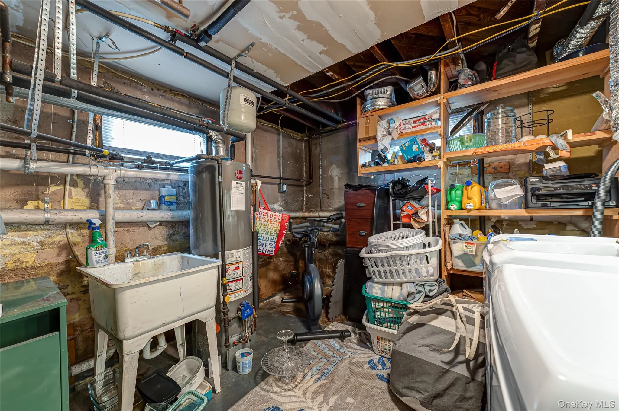 Basement utility area featuring a window, exposed piping, a utility sink, a water heater, and built-in wood shelving Basement utility area featuring a window, exposed piping, a utility sink, a water heater, and built-in wood shelving