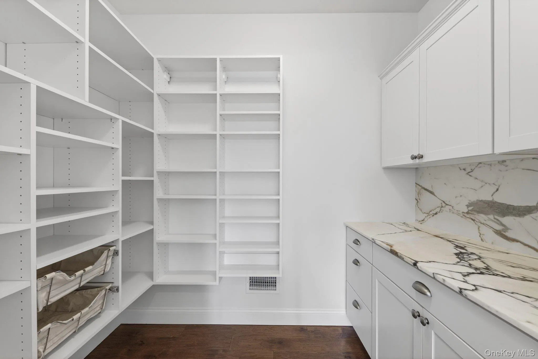 Spacious pantry featuring extensive white shelving, rich wood-finish flooring, white cabinetry, and a stone countertop with matching backsplash Spacious pantry featuring extensive white shelving, rich wood-finish flooring, white cabinetry, and a stone countertop with matching backsplash