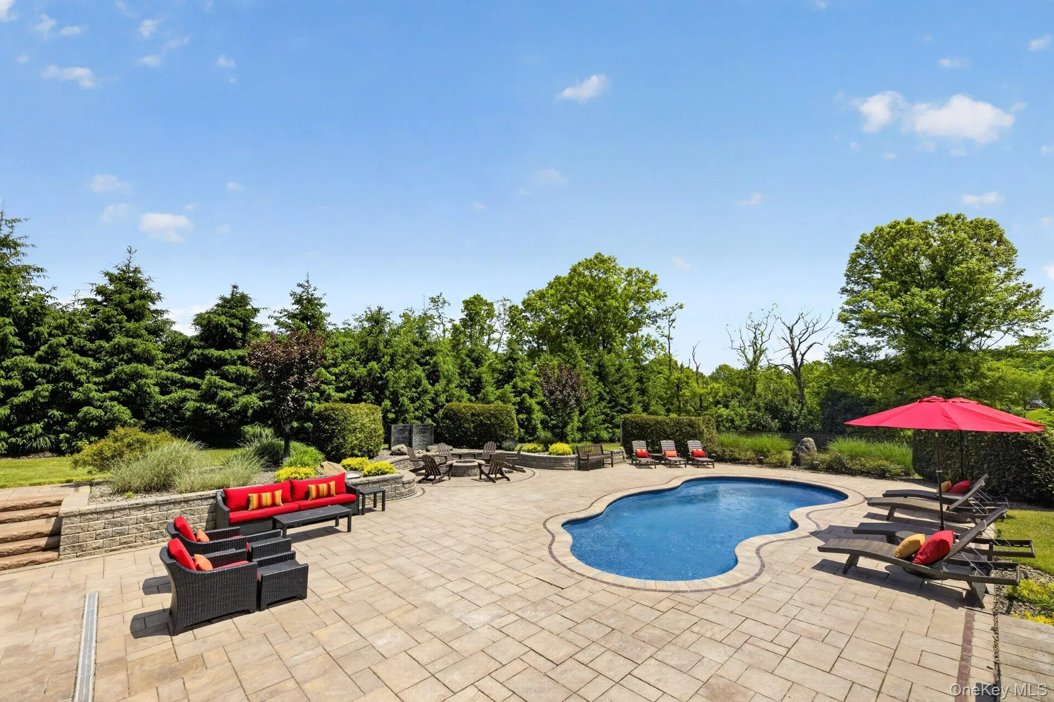 Expansive paver patio featuring a freeform swimming pool, stone retaining walls, and mature perimeter landscaping Expansive paver patio featuring a freeform swimming pool, stone retaining walls, and mature perimeter landscaping