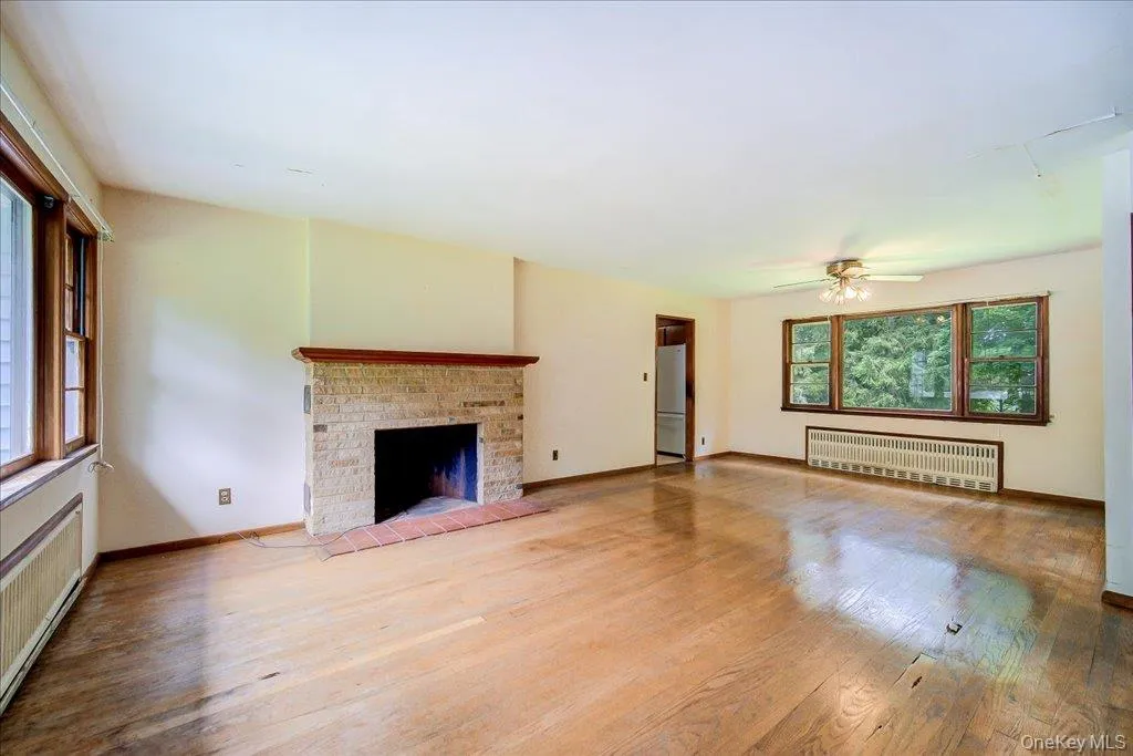 Spacious living area featuring wood-finish flooring, a stone fireplace with a brick hearth, multiple windows with wood trim, and a ceiling fan Spacious living area featuring wood-finish flooring, a stone fireplace with a brick hearth, multiple windows with wood trim, and a ceiling fan