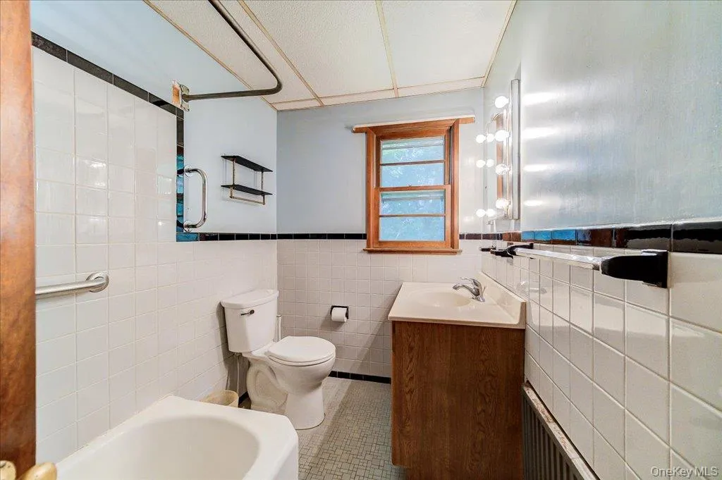 Bathroom featuring white square tile walls with a black trim accent, a white built-in tub, a wood-finish vanity with an integrated sink, and a framed window Bathroom featuring white square tile walls with a black trim accent, a white built-in tub, a wood-finish vanity with an integrated sink, and a framed window