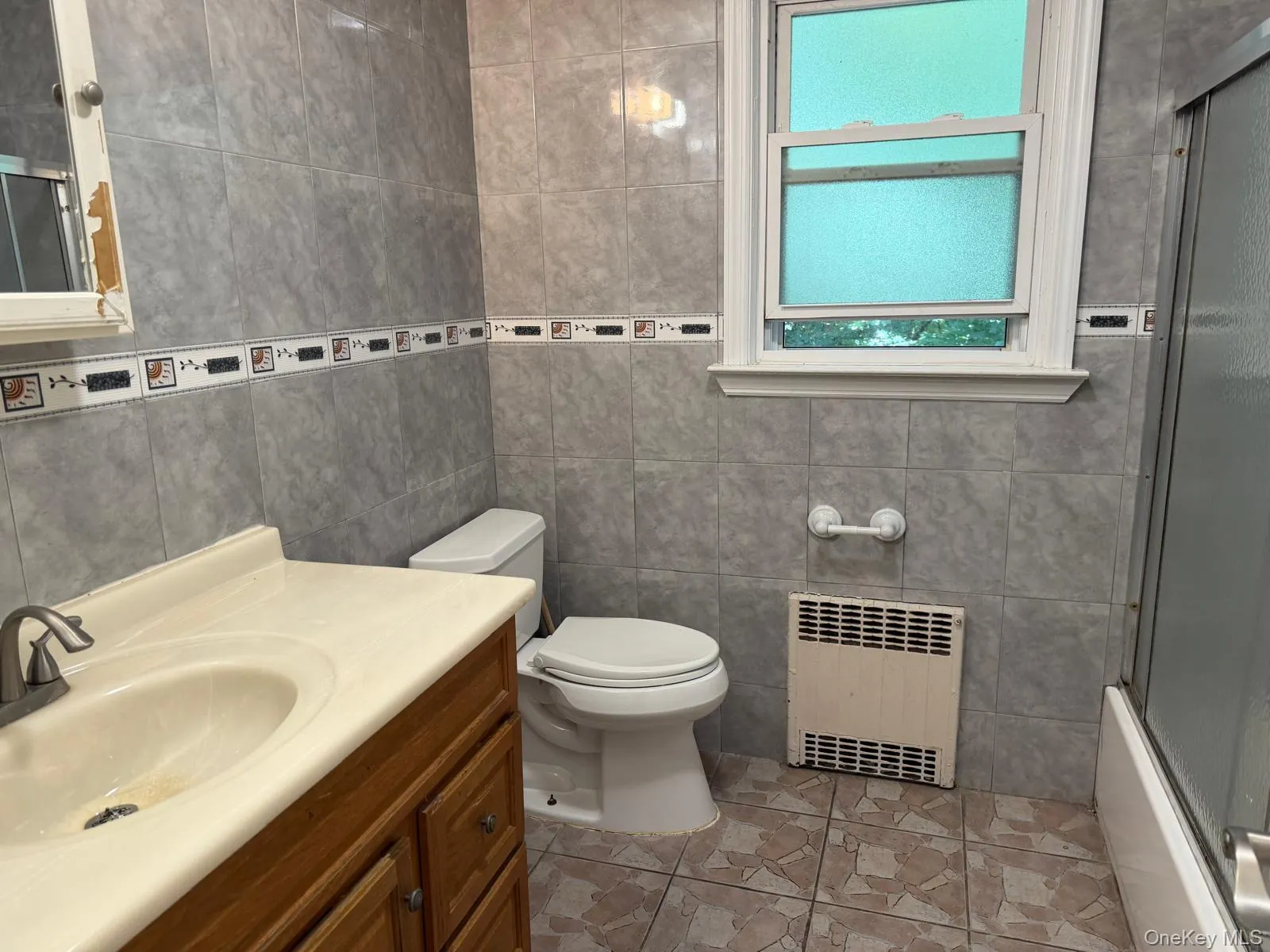 Bathroom featuring full-height gray wall tiling with a decorative border, a wood-finish vanity with an integrated sink, and a single-hung window with privacy glass Bathroom featuring full-height gray wall tiling with a decorative border, a wood-finish vanity with an integrated sink, and a single-hung window with privacy glass