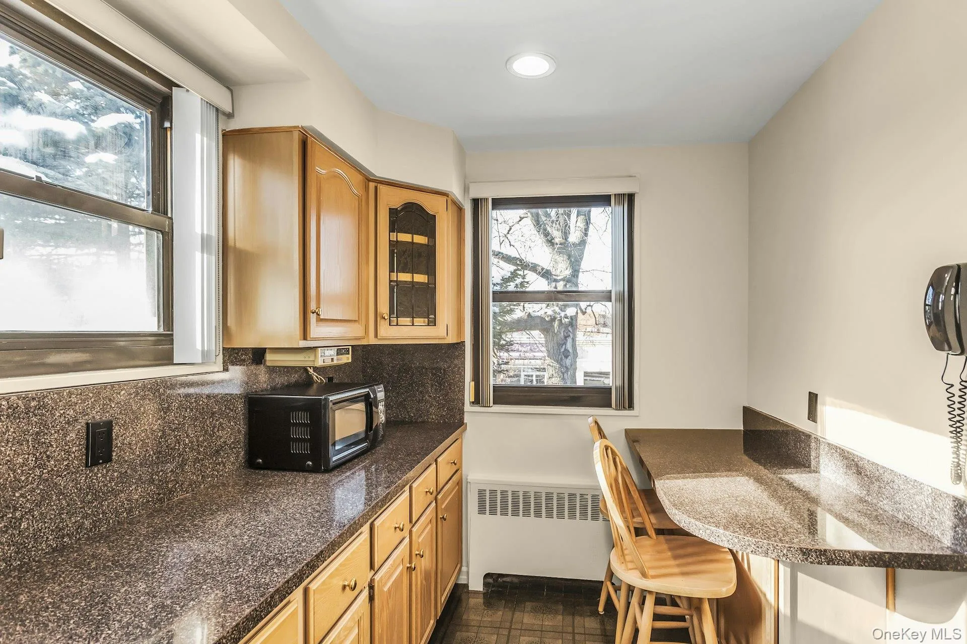Kitchen with a breakfast bar area, dark stone counters, glass insert cabinets, brown cabinetry, and decorative backsplash Kitchen with a breakfast bar area, dark stone counters, glass insert cabinets, brown cabinetry, and decorative backsplash