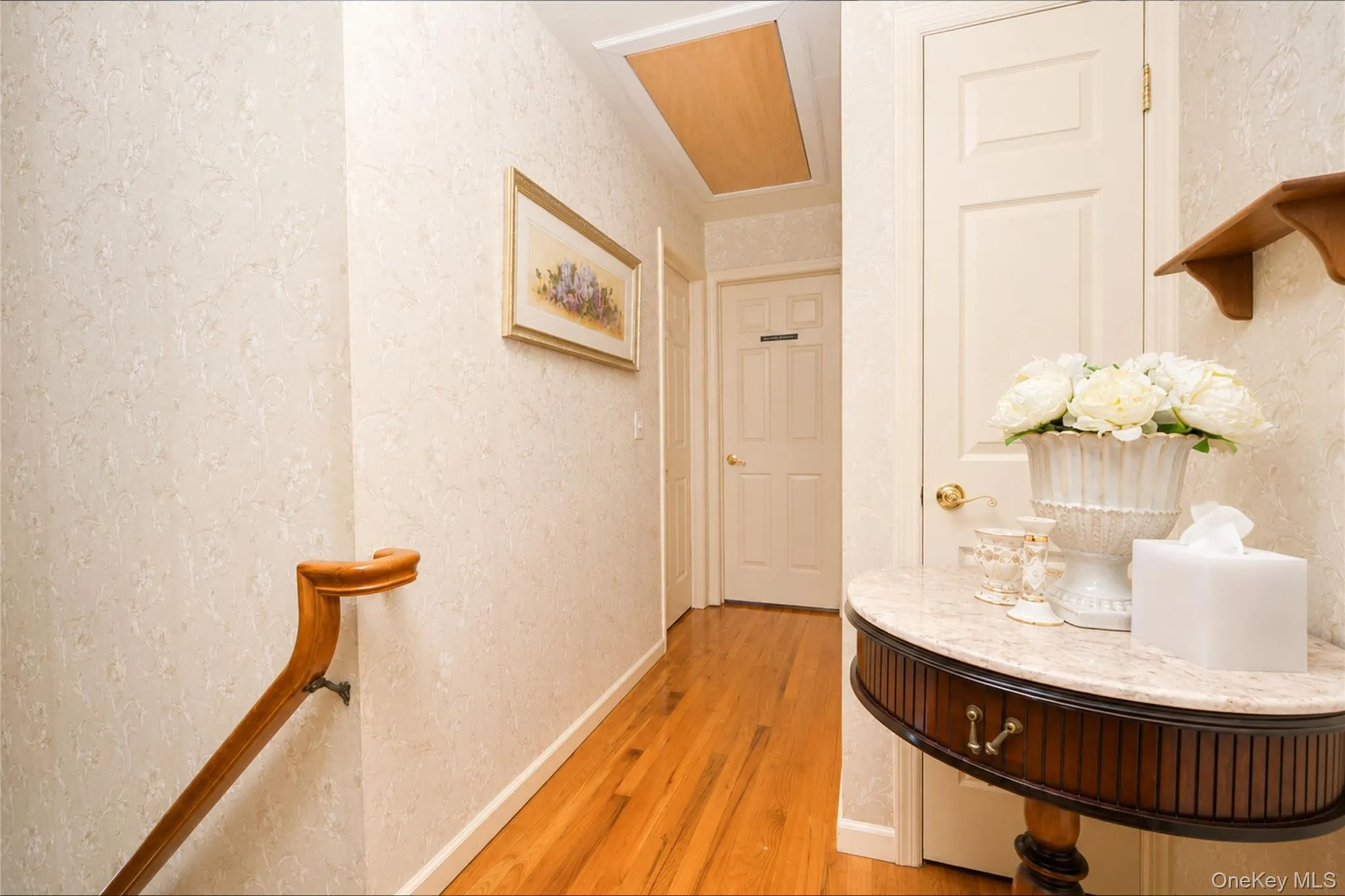Hallway featuring warm wood flooring, textured wallpaper, and a solid wood handrail with decorative carving Hallway featuring warm wood flooring, textured wallpaper, and a solid wood handrail with decorative carving