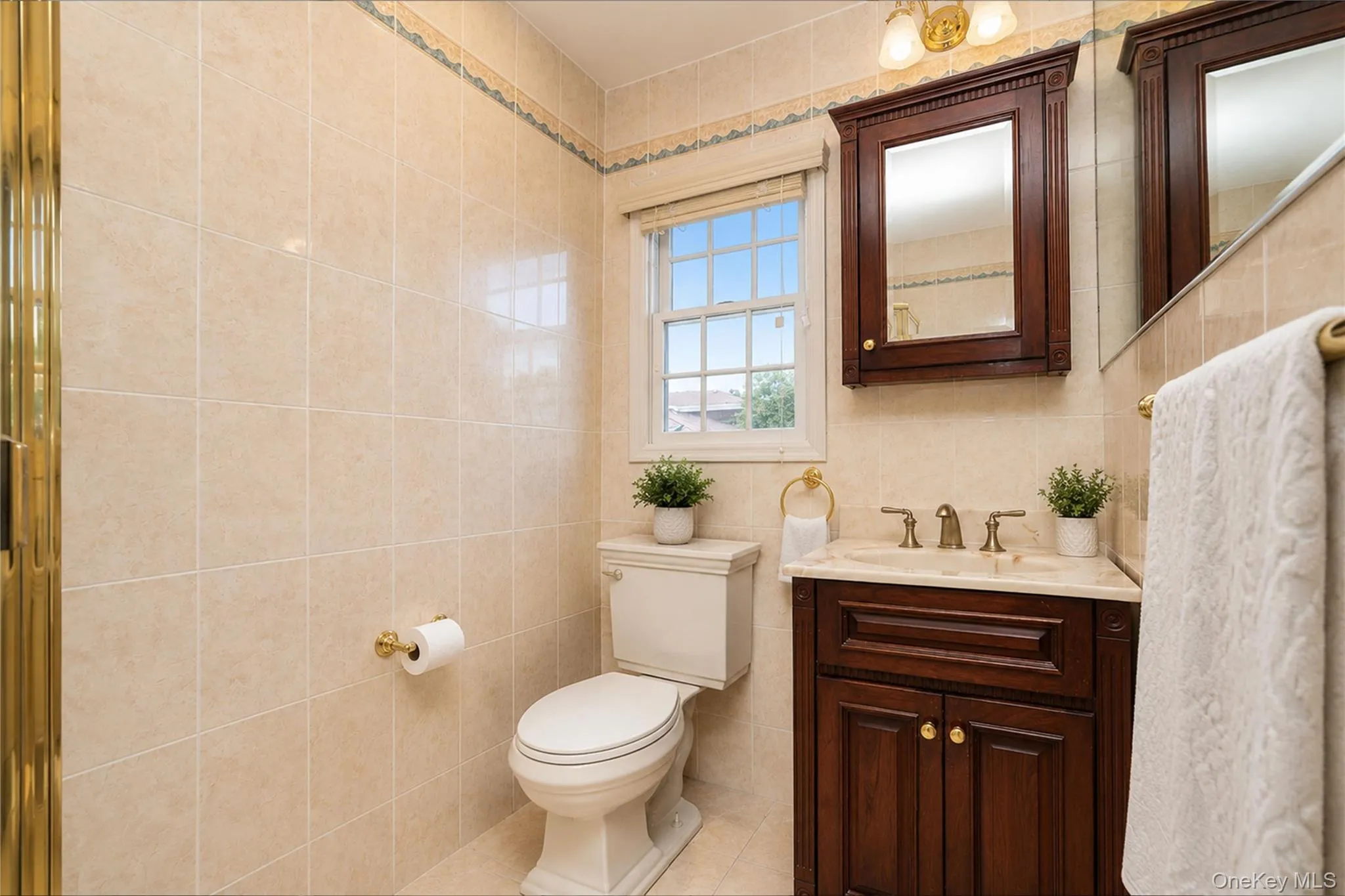 Bathroom featuring a window with privacy blind, full wall and floor tiling, a dark wood-finish vanity with an integrated sink, a matching wall-mounted medicine cabinet, and brass-tone fixtures Bathroom featuring a window with privacy blind, full wall and floor tiling, a dark wood-finish vanity with an integrated sink, a matching wall-mounted medicine cabinet, and brass-tone fixtures