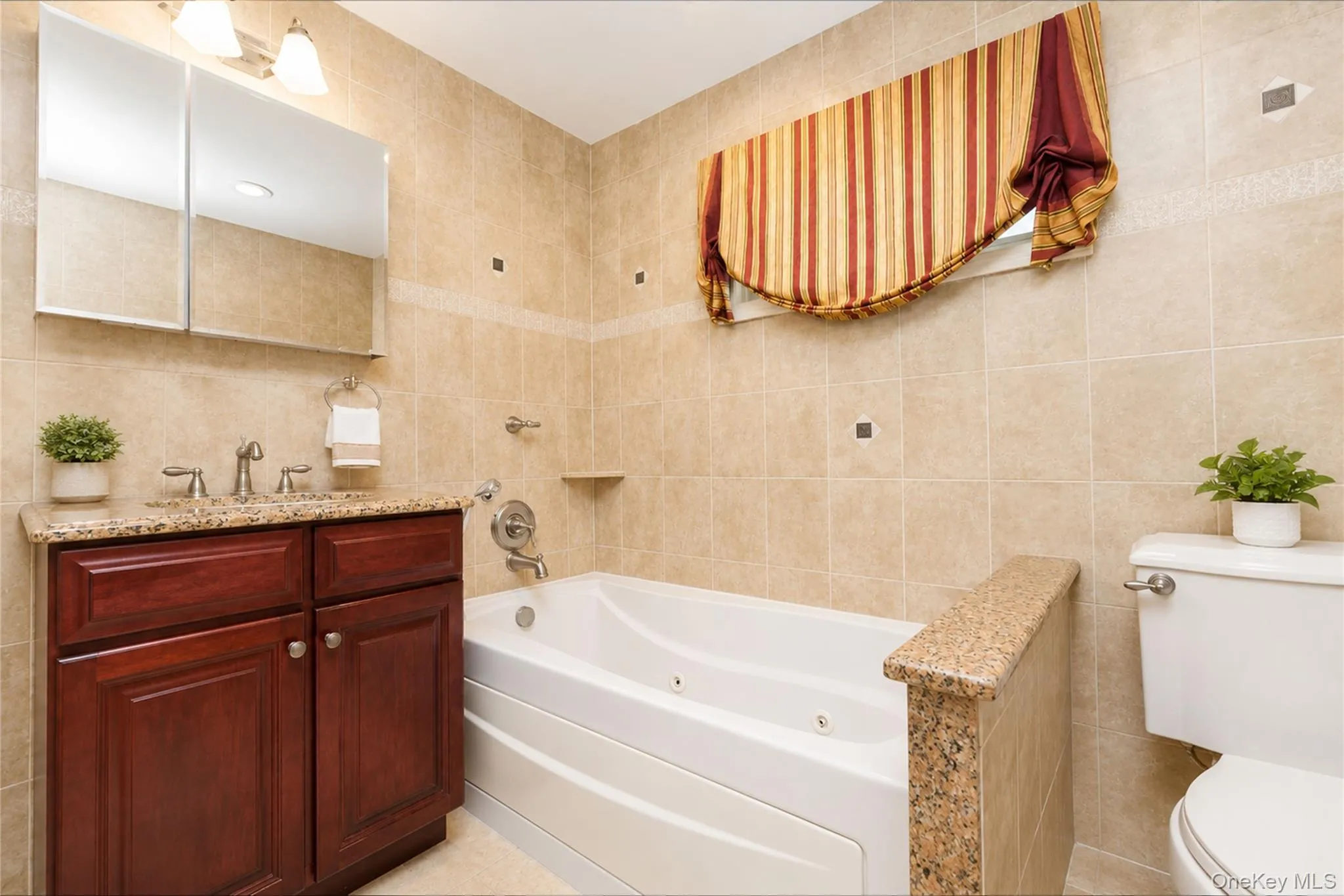 Bathroom featuring a built-in jetted tub, a vanity with a stone countertop and cherry wood-finish cabinetry, a wall-mounted medicine cabinet, and tiled walls with decorative accents Bathroom featuring a built-in jetted tub, a vanity with a stone countertop and cherry wood-finish cabinetry, a wall-mounted medicine cabinet, and tiled walls with decorative accents