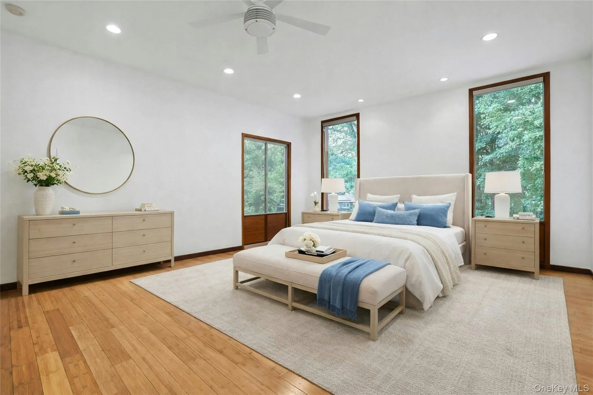 The expansive primary suite is a peaceful retreat with oversized windows framing tranquil wooded views, beautiful hardwood flooring, recessed lighting, and abundant space for a king-sized bed. (Digitally staged.) The expansive primary suite is a peaceful retreat with oversized windows framing tranquil wooded views, beautiful hardwood flooring, recessed lighting, and abundant space for a king-sized bed. (Digitally staged.)