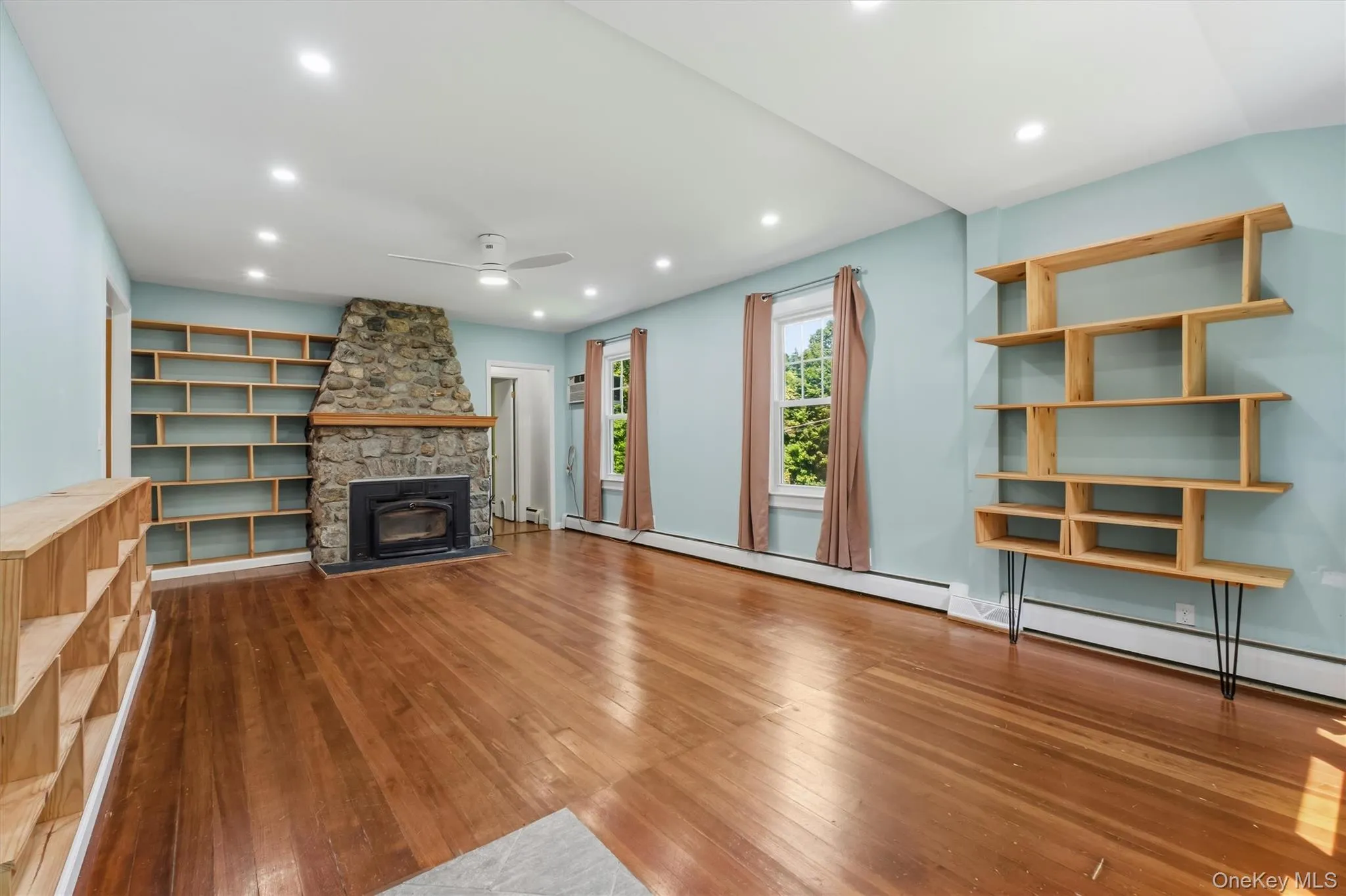 Living area featuring a stone fireplace with a wood mantle, wood-finish flooring, built-in shelving, recessed lighting, and a ceiling fan Living area featuring a stone fireplace with a wood mantle, wood-finish flooring, built-in shelving, recessed lighting, and a ceiling fan