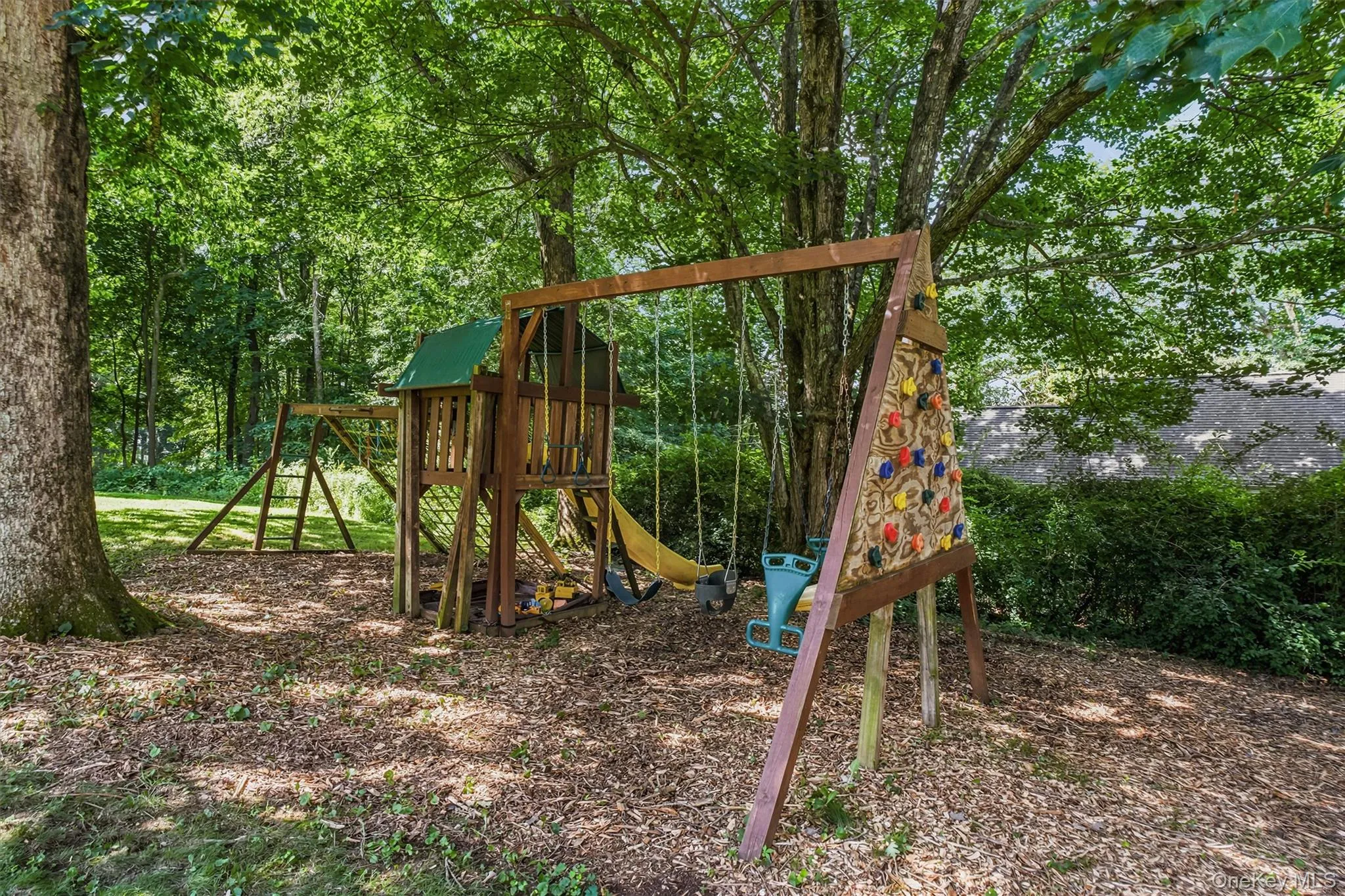 Wood-toned playset with a green roof, integrated slide, and swing set, set upon a wood chip ground cover Wood-toned playset with a green roof, integrated slide, and swing set, set upon a wood chip ground cover