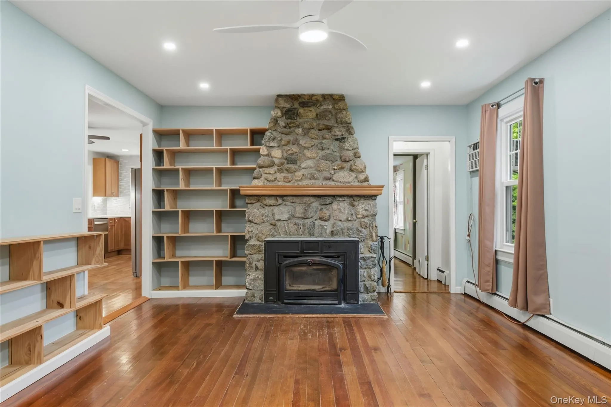 Living area featuring a stone fireplace with a wood mantle, wood-finish flooring, built-in shelving, recessed lighting, and a ceiling fan Living area featuring a stone fireplace with a wood mantle, wood-finish flooring, built-in shelving, recessed lighting, and a ceiling fan