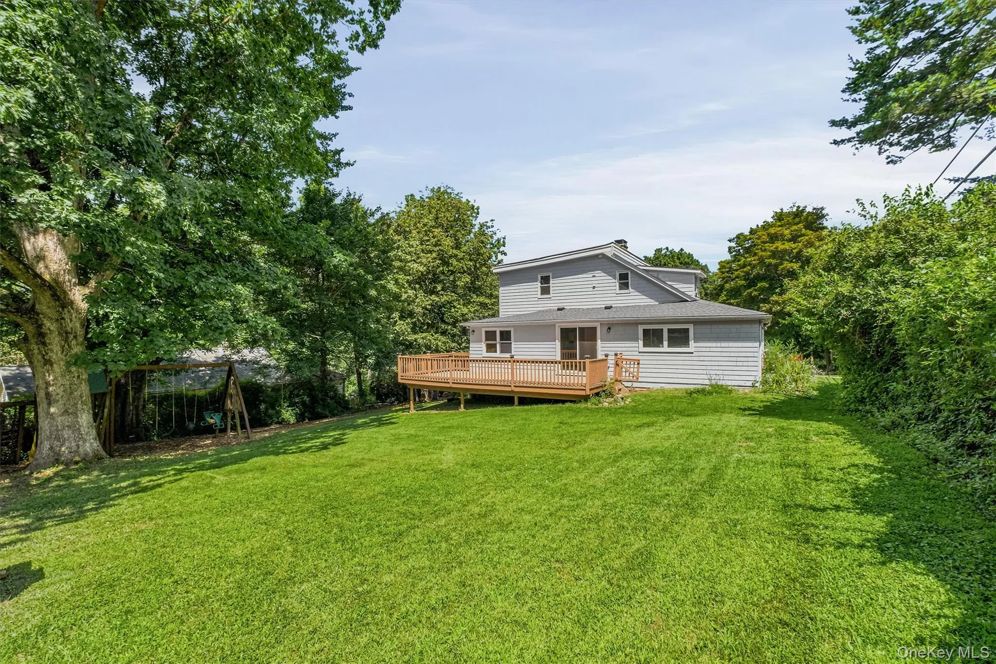 Expansive rear yard featuring a large wooden deck, dual sliding glass doors, a grey siding exterior, a multi-level roofline, and mature perimeter trees Expansive rear yard featuring a large wooden deck, dual sliding glass doors, a grey siding exterior, a multi-level roofline, and mature perimeter trees