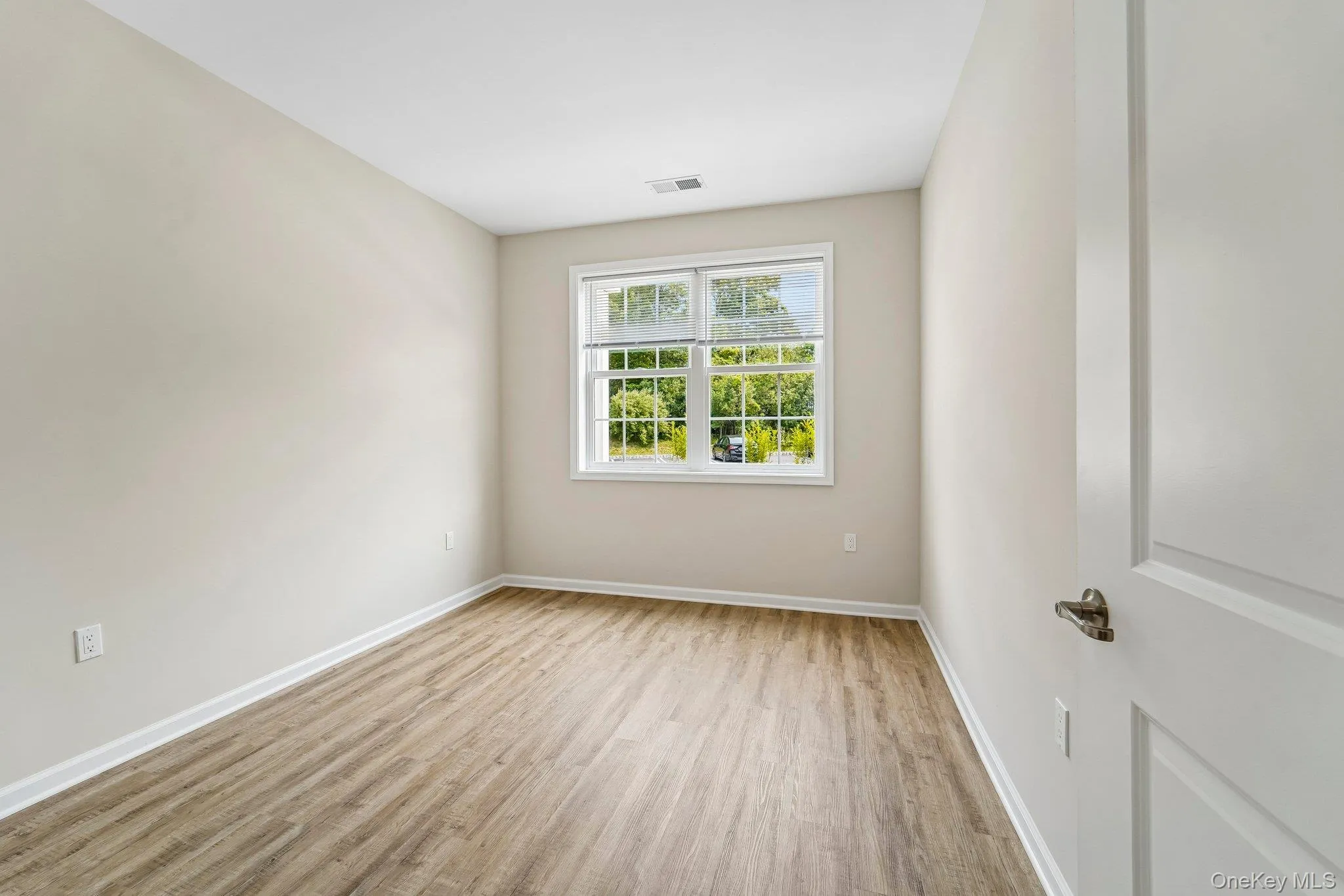 Light-filled room featuring wood-finish flooring, neutral wall paint, white trim, and a white interior door with bronze hardware Light-filled room featuring wood-finish flooring, neutral wall paint, white trim, and a white interior door with bronze hardware