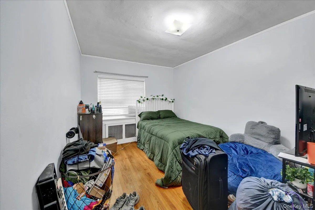 Bedroom featuring wood-finish flooring, light gray walls, a window with blinds, and a built-in radiator cover Bedroom featuring wood-finish flooring, light gray walls, a window with blinds, and a built-in radiator cover