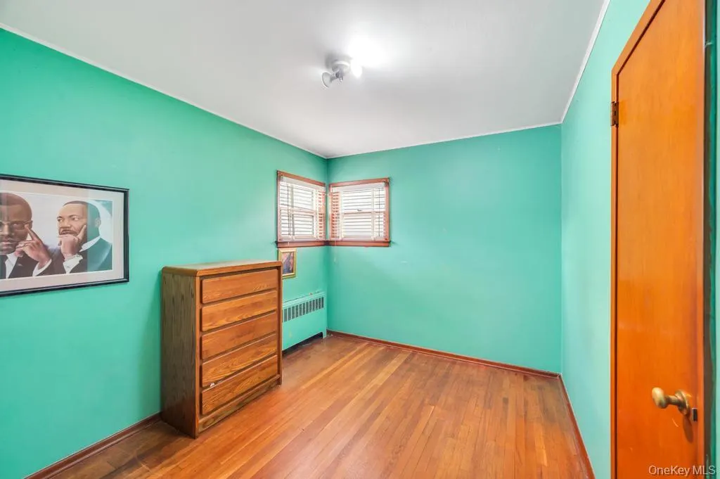 Interior room featuring wood-finish flooring, two windows with blinds, a wall-mounted radiator, and a flush-mount ceiling light Interior room featuring wood-finish flooring, two windows with blinds, a wall-mounted radiator, and a flush-mount ceiling light