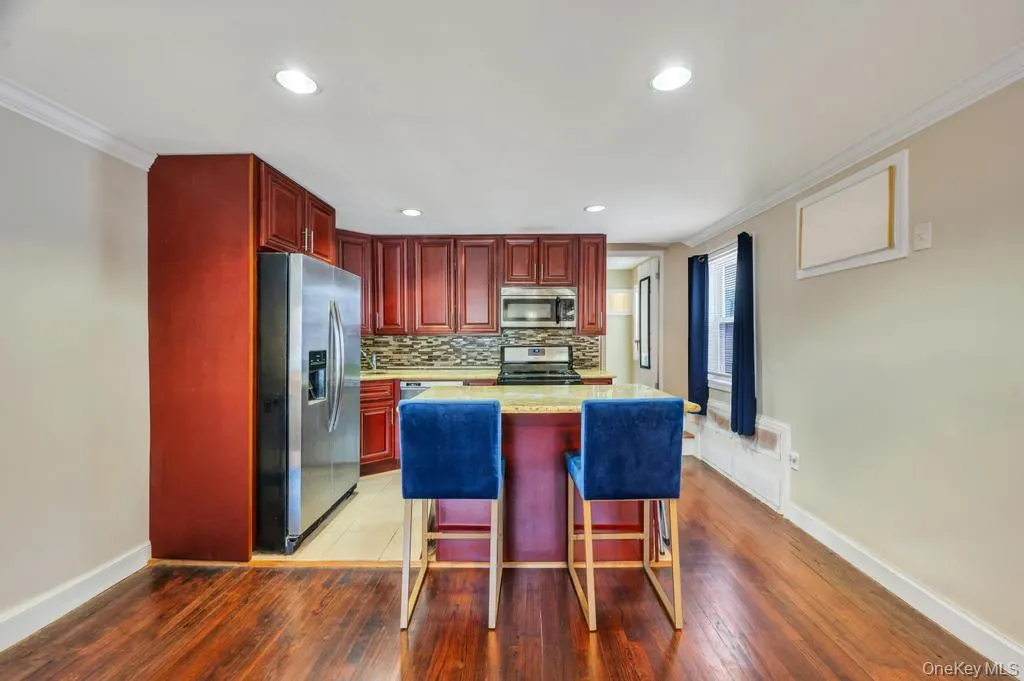 Open-concept kitchen featuring wood-finish cabinetry, stainless steel appliances, a multi-tone tile backsplash, and a central island with a light-colored countertop Open-concept kitchen featuring wood-finish cabinetry, stainless steel appliances, a multi-tone tile backsplash, and a central island with a light-colored countertop