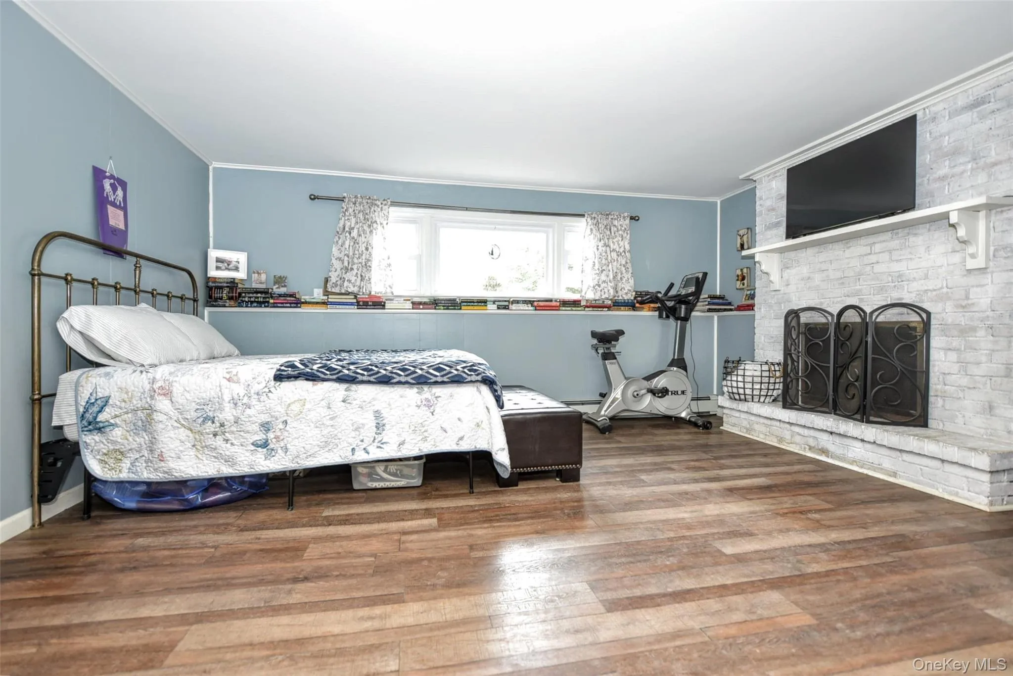 Room featuring wood-finish flooring, light blue walls, and a white brick-style fireplace with a mantel Room featuring wood-finish flooring, light blue walls, and a white brick-style fireplace with a mantel