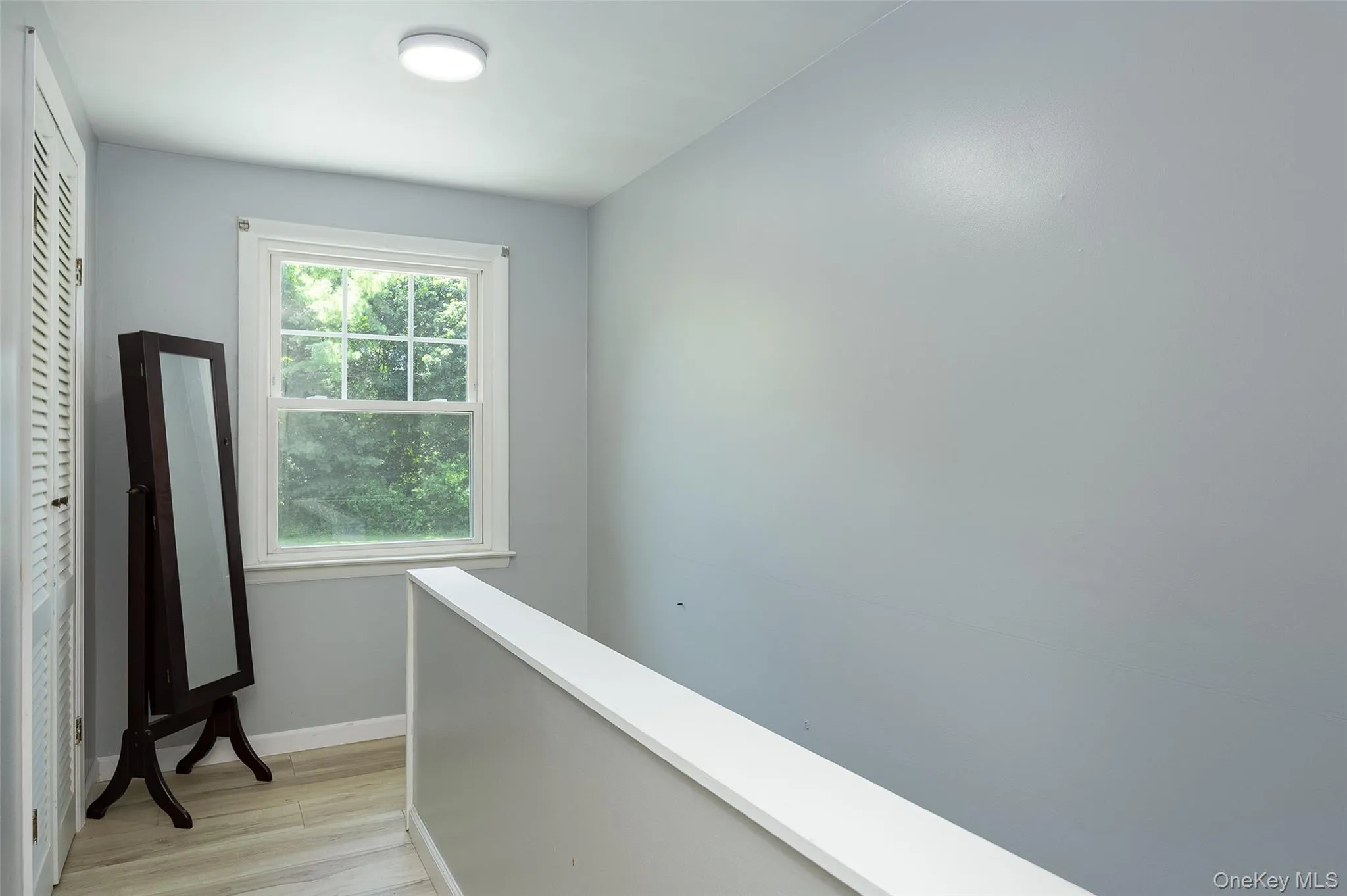 Bright interior space featuring a window with exterior tree scenery, wood-finish flooring, a contemporary flush-mount ceiling light, and a white balustrade Bright interior space featuring a window with exterior tree scenery, wood-finish flooring, a contemporary flush-mount ceiling light, and a white balustrade