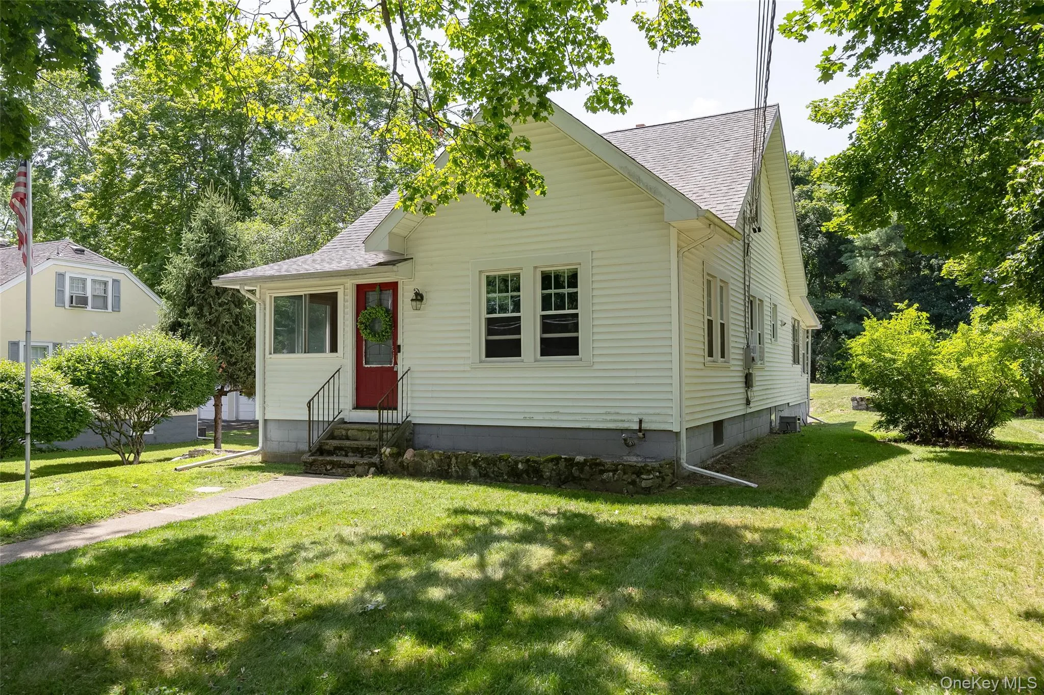 White siding residence featuring a red entry door, an enclosed porch, a stone foundation, a gabled roofline, and a mature tree canopy White siding residence featuring a red entry door, an enclosed porch, a stone foundation, a gabled roofline, and a mature tree canopy