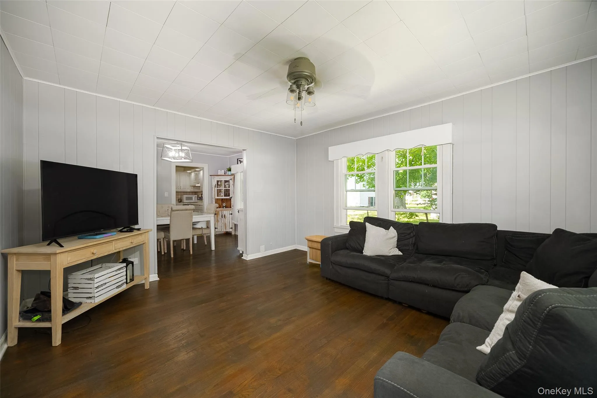 Spacious living area featuring wood-finish flooring, light gray vertical paneling, and a white paneled ceiling Spacious living area featuring wood-finish flooring, light gray vertical paneling, and a white paneled ceiling
