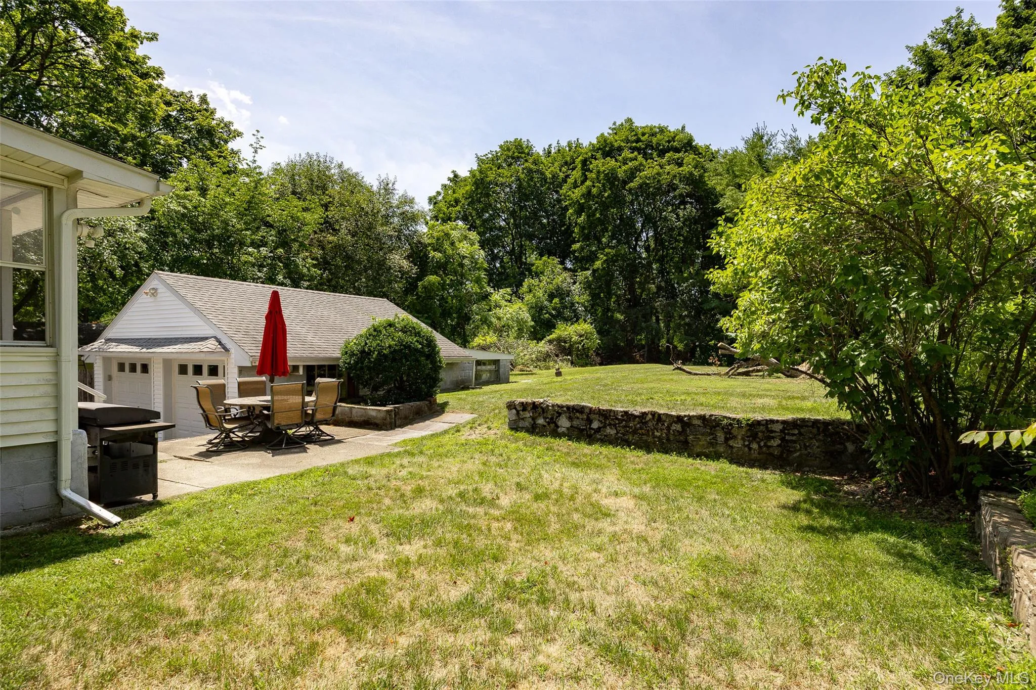 Expansive backyard featuring a stone retaining wall, mature trees, and a concrete patio Expansive backyard featuring a stone retaining wall, mature trees, and a concrete patio