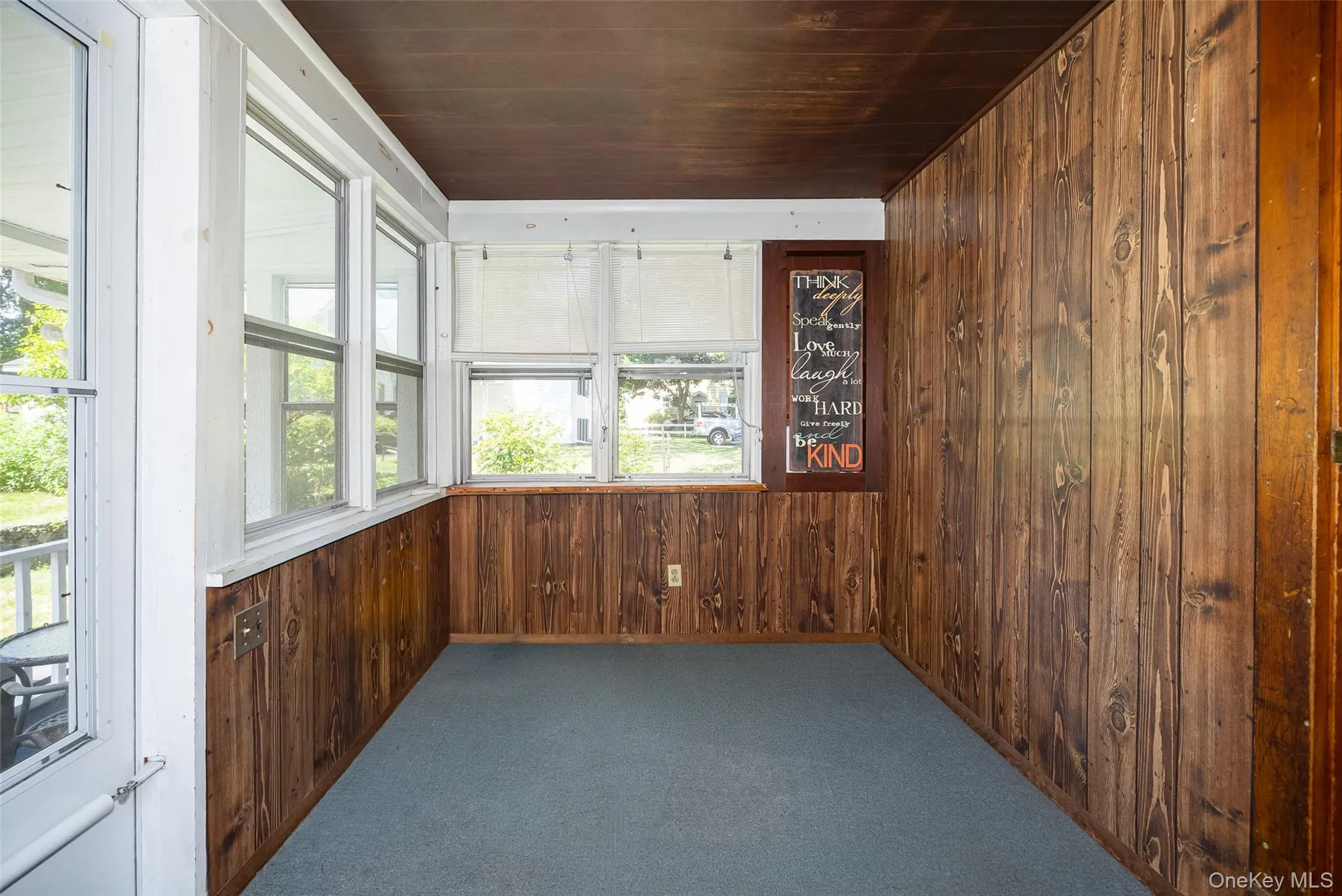 Enclosed porch featuring wood-panel walls, multiple windows providing natural illumination, and a blue carpeted floor Enclosed porch featuring wood-panel walls, multiple windows providing natural illumination, and a blue carpeted floor