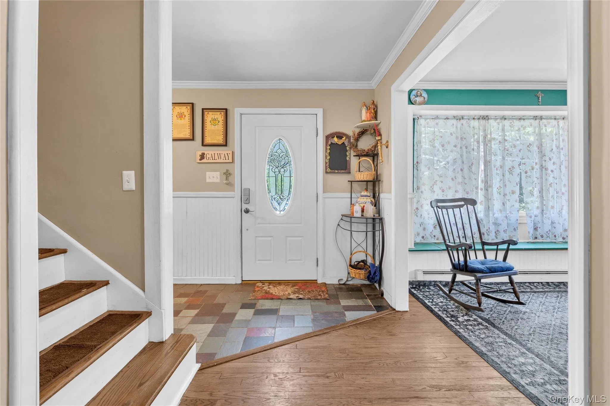 Entryway featuring a white paneled door with an oval stained-glass insert, multi-toned tile flooring, and white wainscoting Entryway featuring a white paneled door with an oval stained-glass insert, multi-toned tile flooring, and white wainscoting