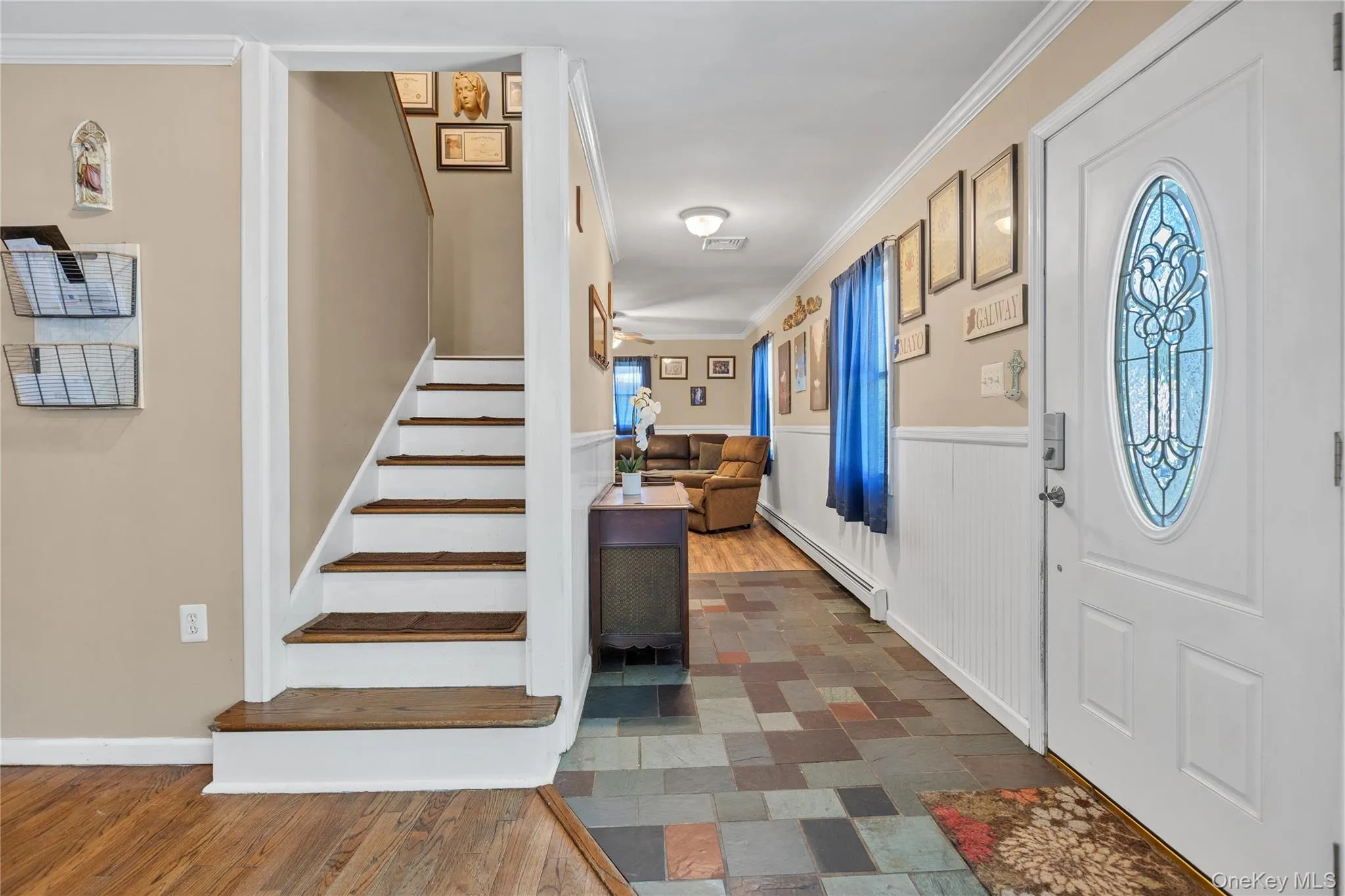 Entryway featuring a white paneled door with an oval stained-glass insert, multi-toned slate-style flooring, white wainscoting, and a staircase with wood-finish treads Entryway featuring a white paneled door with an oval stained-glass insert, multi-toned slate-style flooring, white wainscoting, and a staircase with wood-finish treads