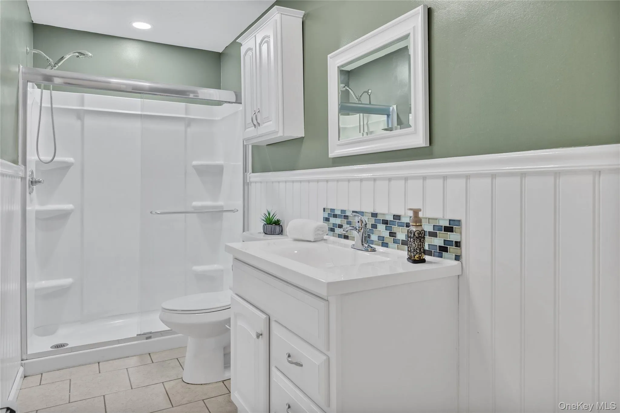 Bathroom featuring a white vanity with integrated sink, patterned tile backsplash, beadboard paneling, and a white shower stall with a sliding glass door Bathroom featuring a white vanity with integrated sink, patterned tile backsplash, beadboard paneling, and a white shower stall with a sliding glass door