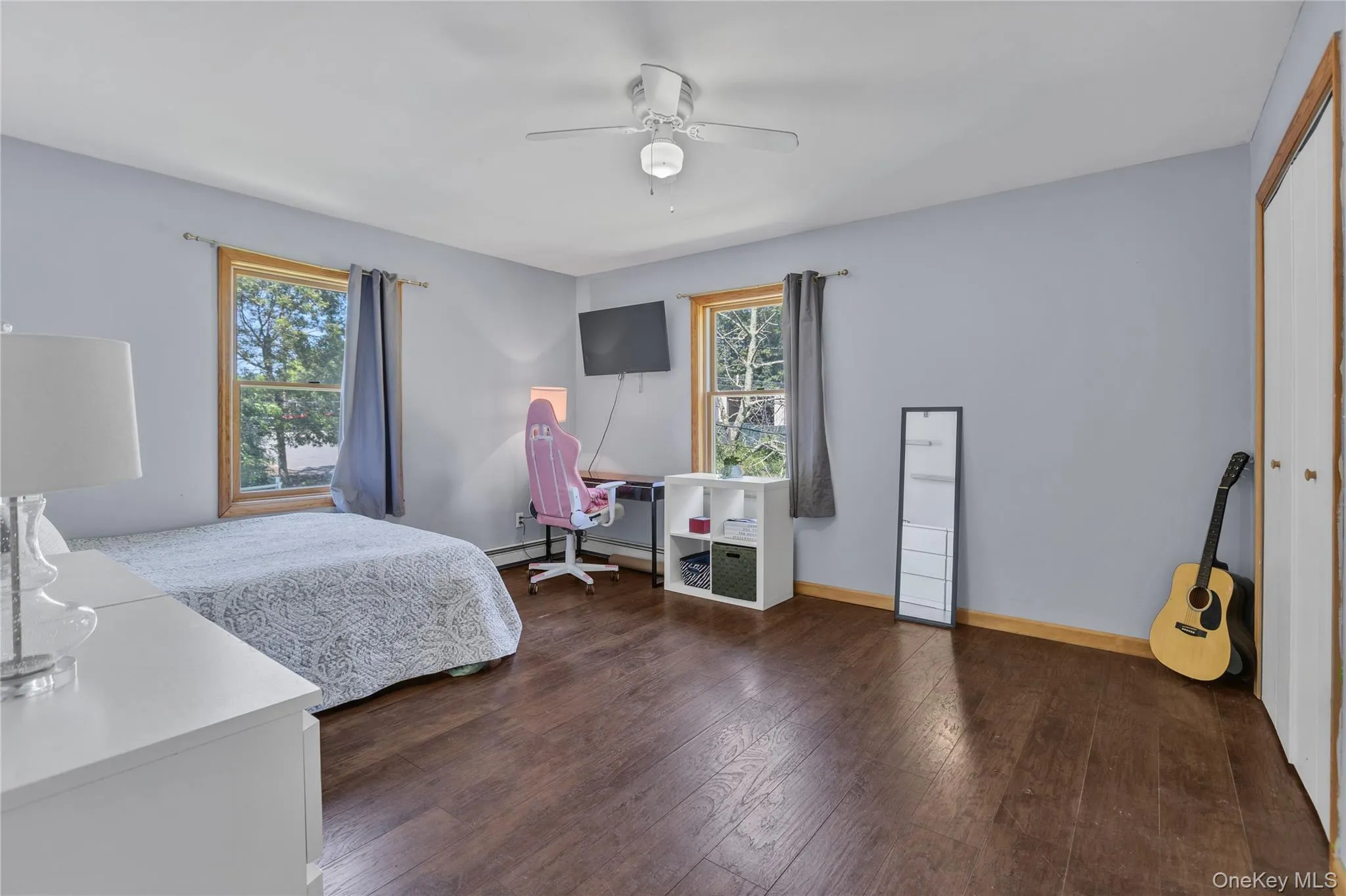Spacious room featuring wood-finish flooring, two windows with wood trim, light gray walls, a white ceiling fan, and a built-in closet with bi-fold doors Spacious room featuring wood-finish flooring, two windows with wood trim, light gray walls, a white ceiling fan, and a built-in closet with bi-fold doors