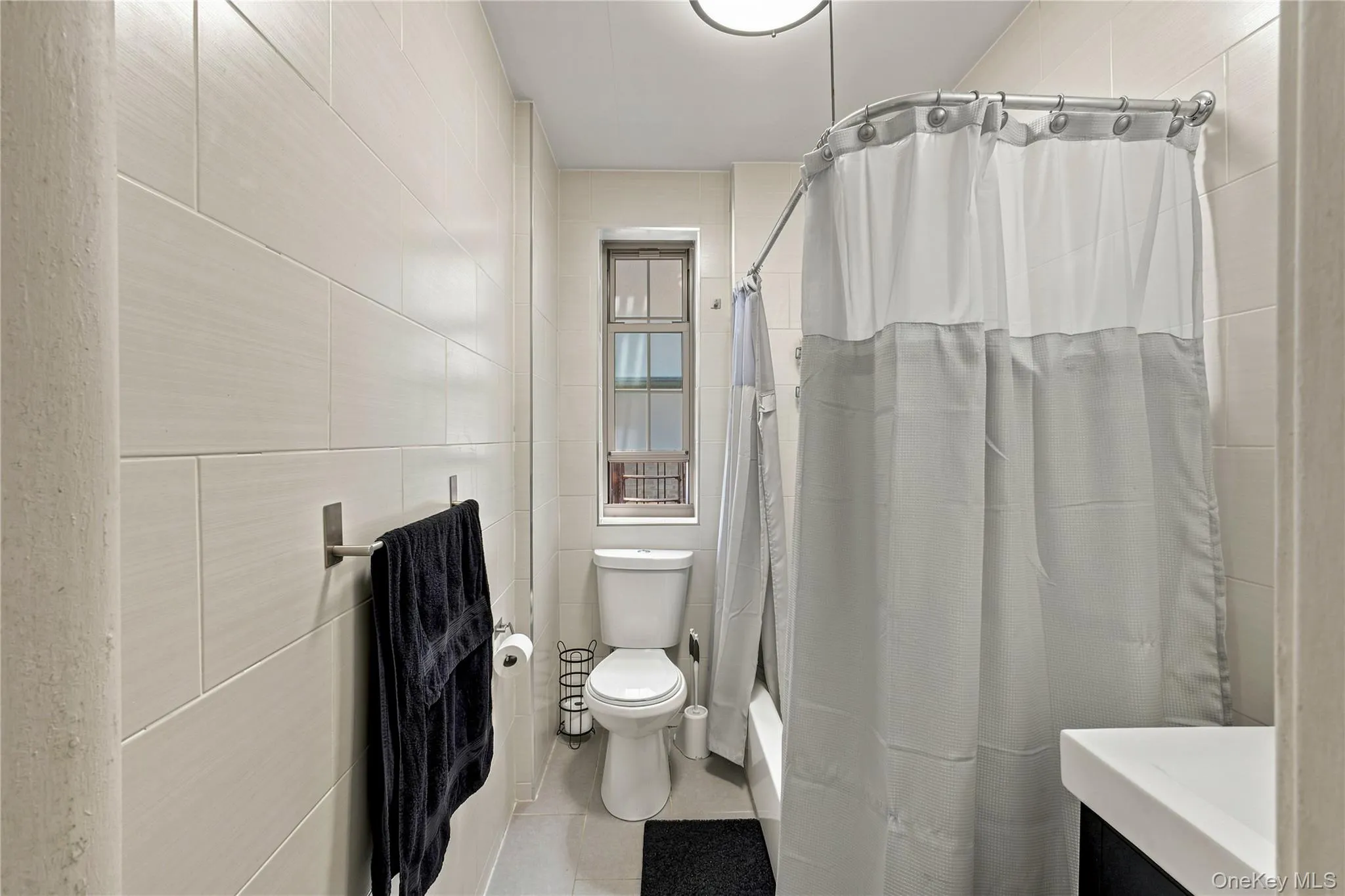 Tiled bathroom featuring a built-in tub with shower, white toilet, and a window with frosted glass Tiled bathroom featuring a built-in tub with shower, white toilet, and a window with frosted glass