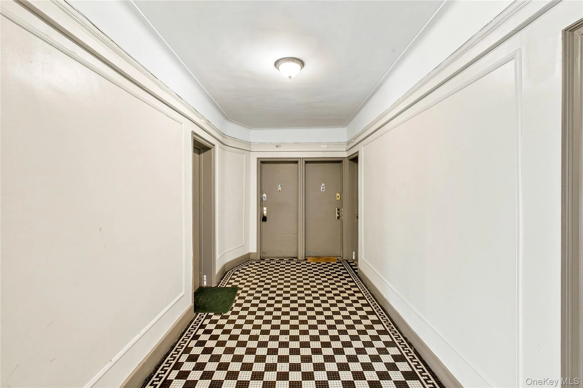 Hallway featuring checkerboard tile flooring with a decorative border, white walls with applied molding details, and a flush mount ceiling light fixture Hallway featuring checkerboard tile flooring with a decorative border, white walls with applied molding details, and a flush mount ceiling light fixture