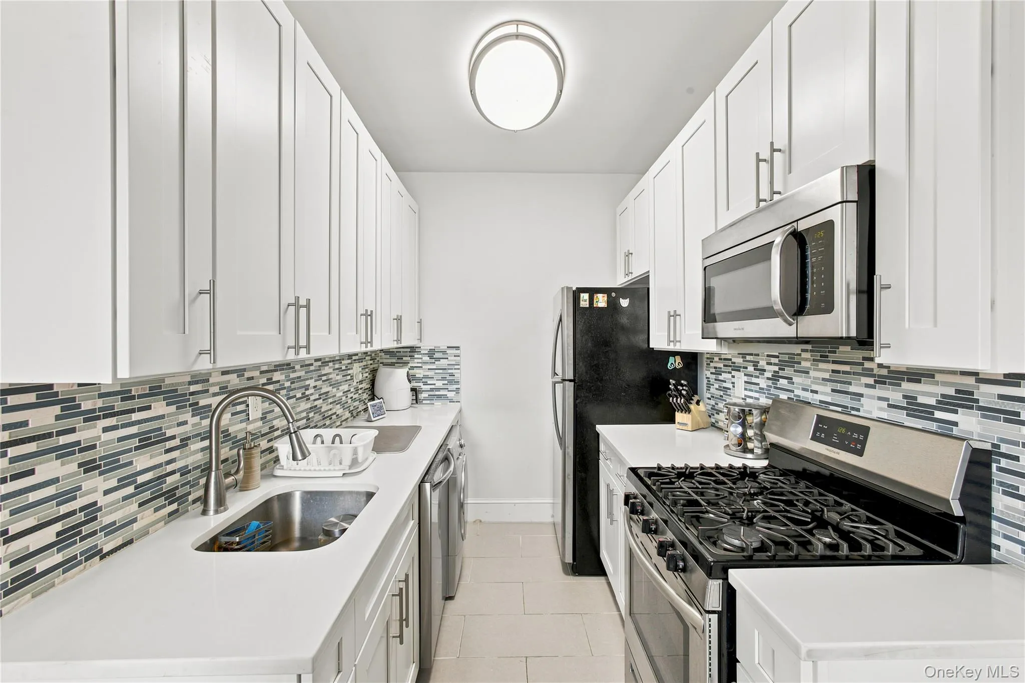 Galley kitchen featuring white cabinetry, quartz countertops, a mosaic tile backsplash, stainless steel appliances, and tile flooring Galley kitchen featuring white cabinetry, quartz countertops, a mosaic tile backsplash, stainless steel appliances, and tile flooring