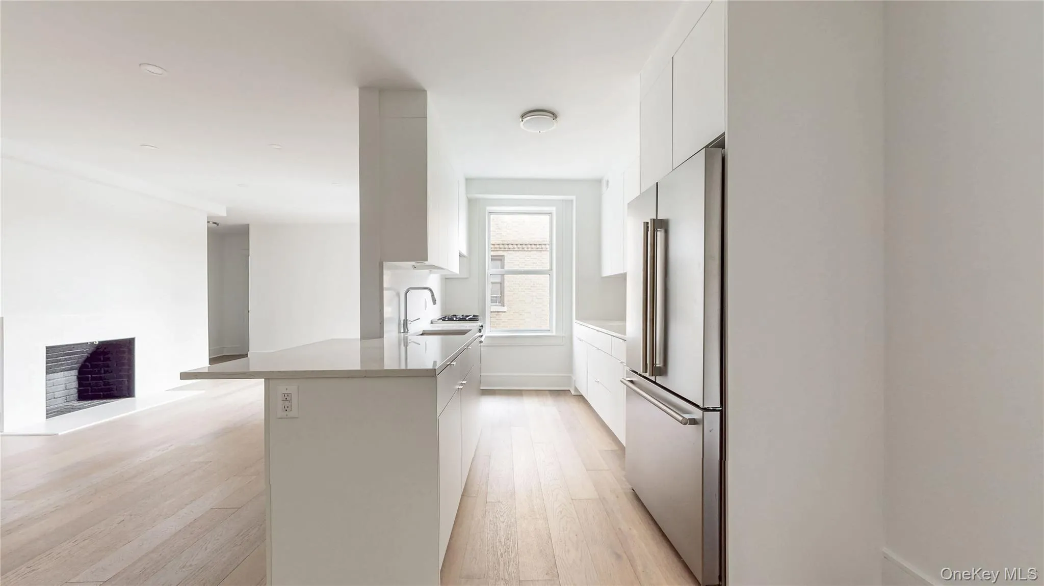 Open-concept kitchen featuring an island with a countertop, stainless steel refrigerator, white cabinetry, integrated sink with a faucet, and light wood-finish flooring Open-concept kitchen featuring an island with a countertop, stainless steel refrigerator, white cabinetry, integrated sink with a faucet, and light wood-finish flooring