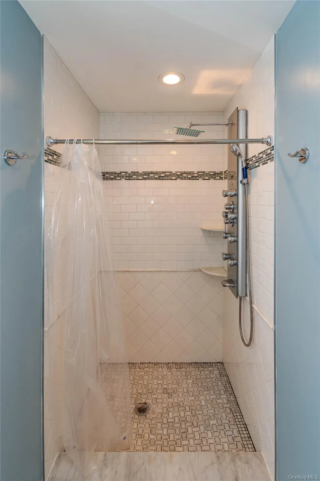 Shower featuring white subway tile walls, a mosaic tile accent band, a multi-function shower panel with an overhead rainfall shower, and a white mosaic tile floor Shower featuring white subway tile walls, a mosaic tile accent band, a multi-function shower panel with an overhead rainfall shower, and a white mosaic tile floor