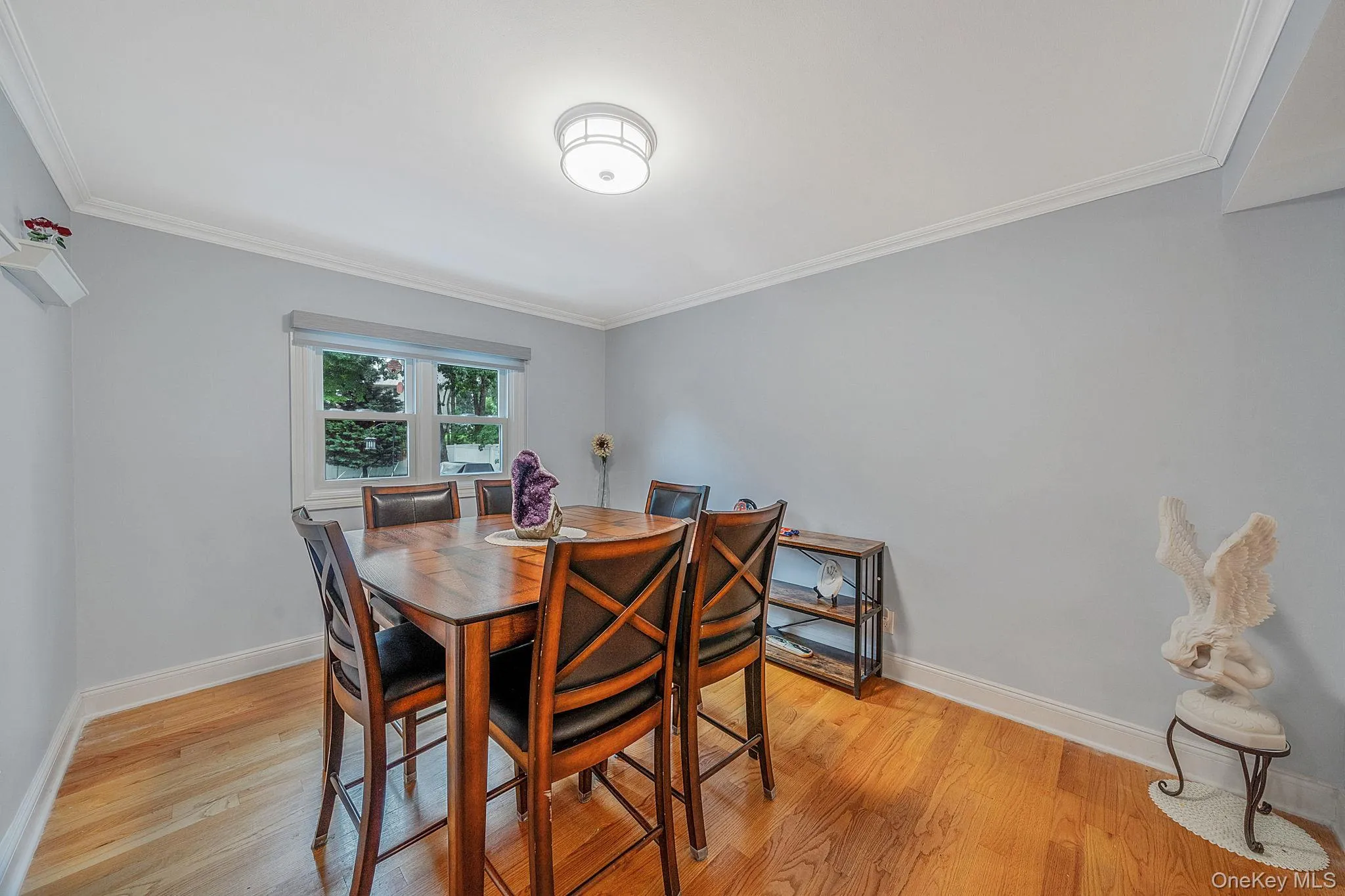 Light-filled ding room featuring hardwood flooring, crown molding, a window with a roller shade, and a flush-mount ceiling light fixture Light-filled ding room featuring hardwood flooring, crown molding, a window with a roller shade, and a flush-mount ceiling light fixture