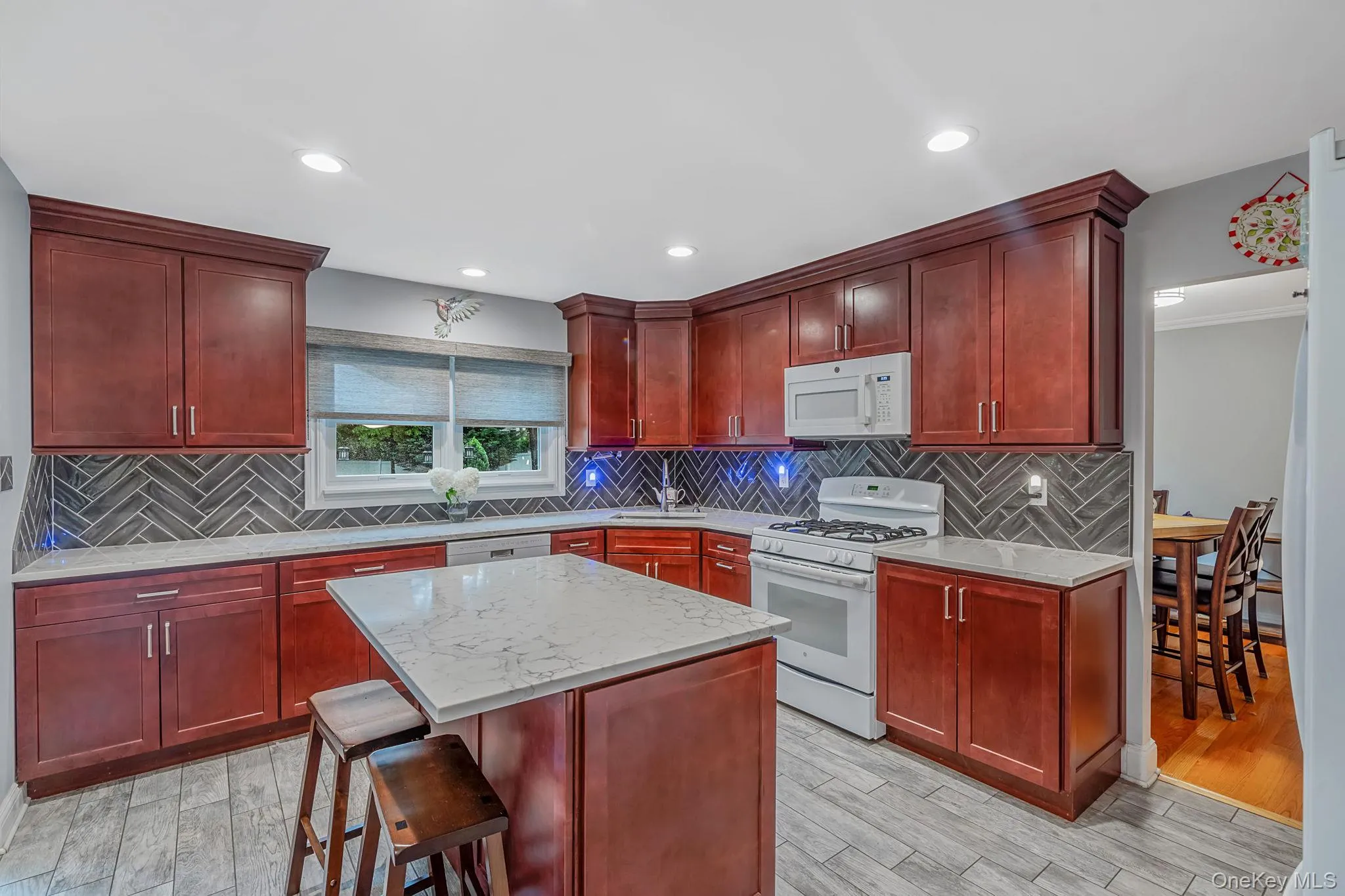 Kitchen featuring rich wood cabinetry, light-toned countertops, a herringbone pattern tile backsplash, white appliances, and recessed lighting Kitchen featuring rich wood cabinetry, light-toned countertops, a herringbone pattern tile backsplash, white appliances, and recessed lighting